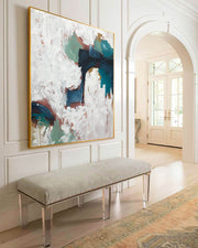 A modern abstract canvas with neutral and dark tones, presented in a floating frame, displayed above a bench in a well-lit room with white walls and a patterned floor.