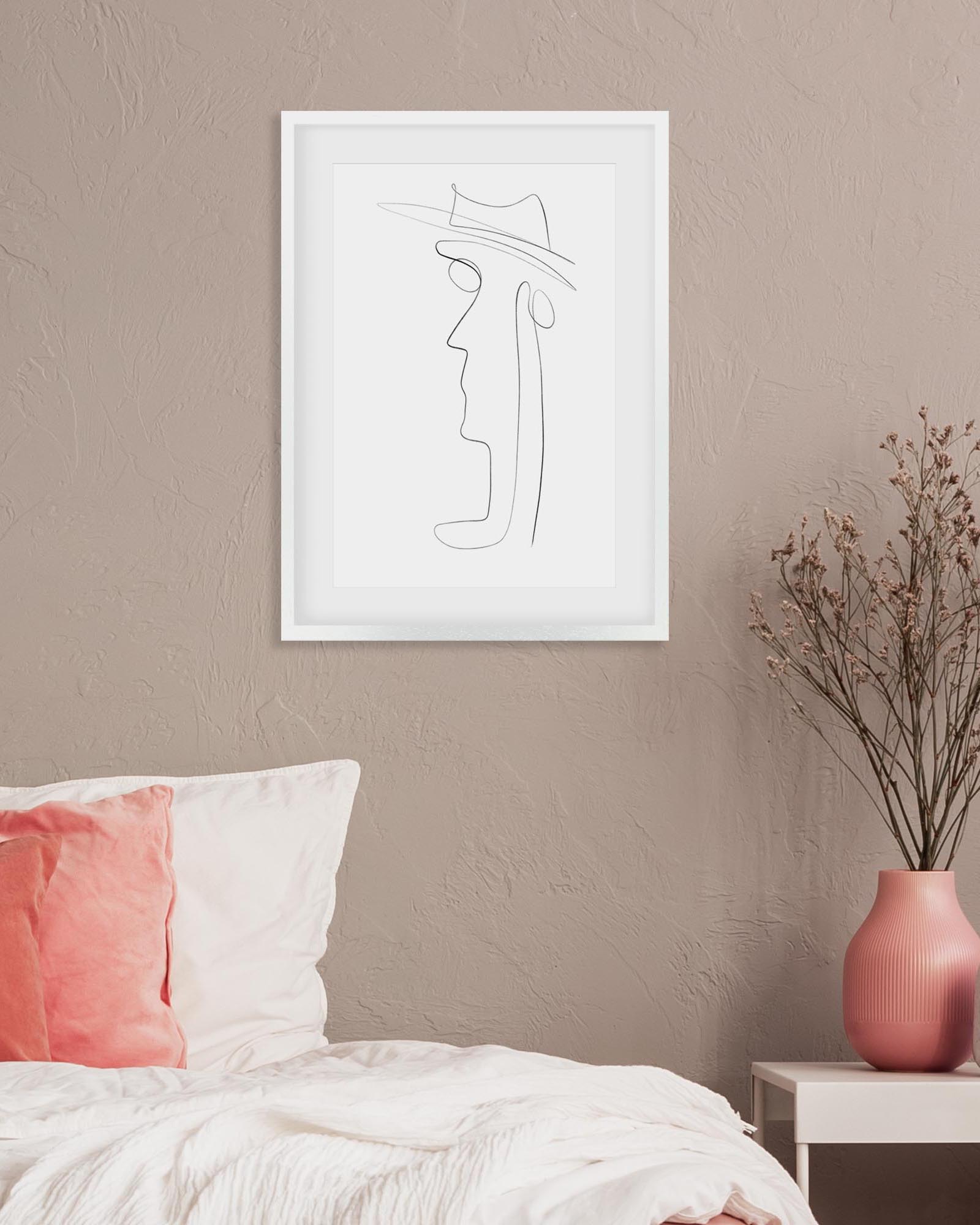 Framed line art of a person wearing a hat on a beige wall above a bed with pink and white bedding.