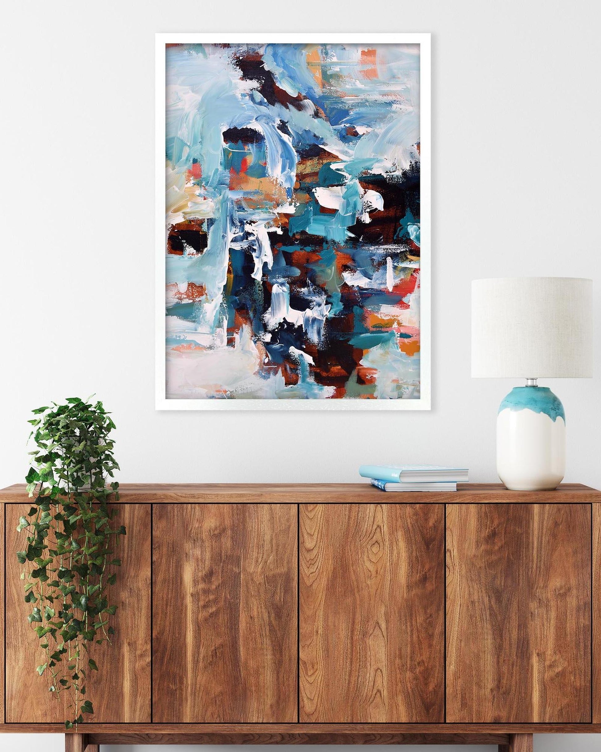 Abstract painting on a wall above a wooden cabinet with a lamp and plant.
