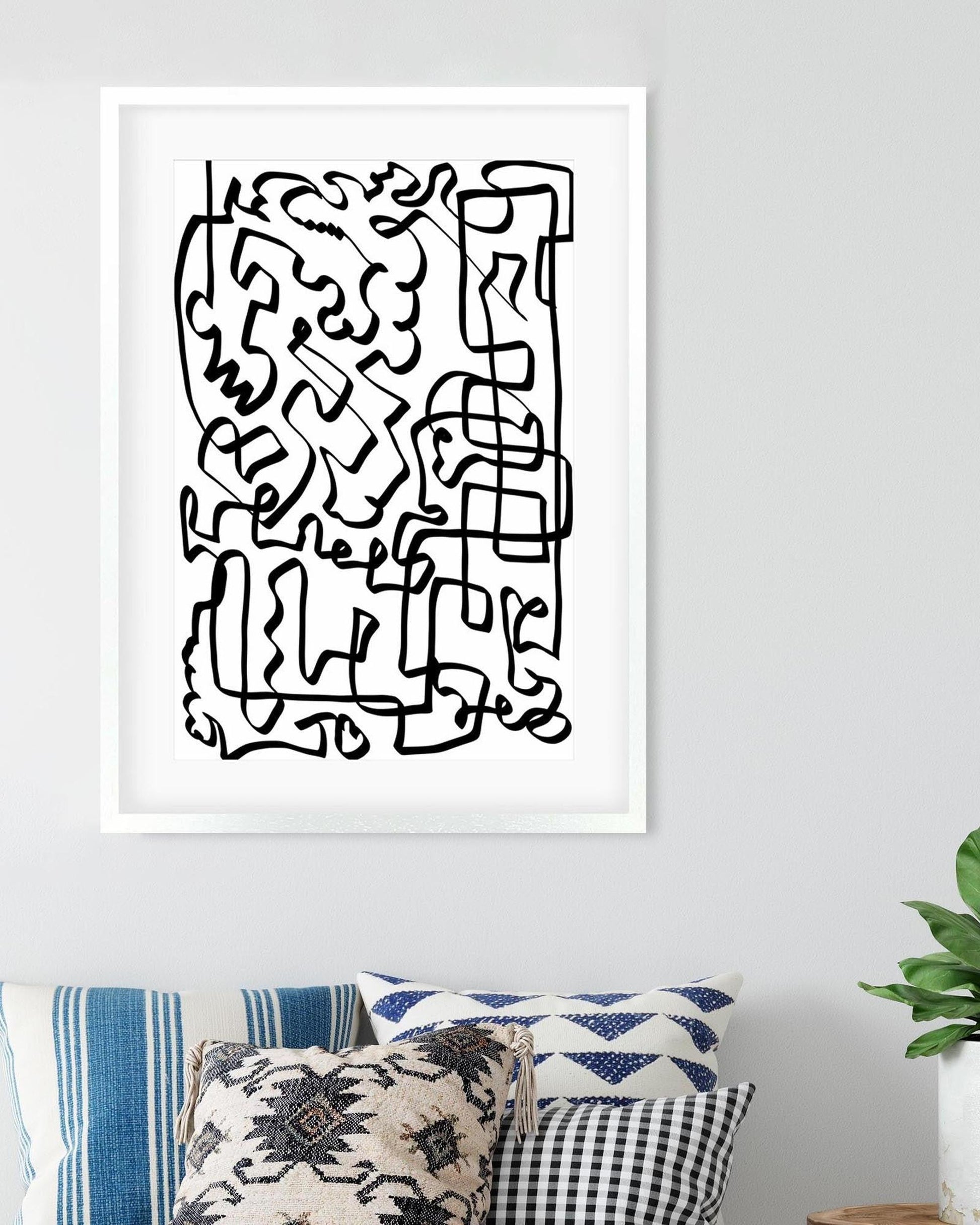 Framed abstract black line art on a white wall with decorative pillows and a plant below.