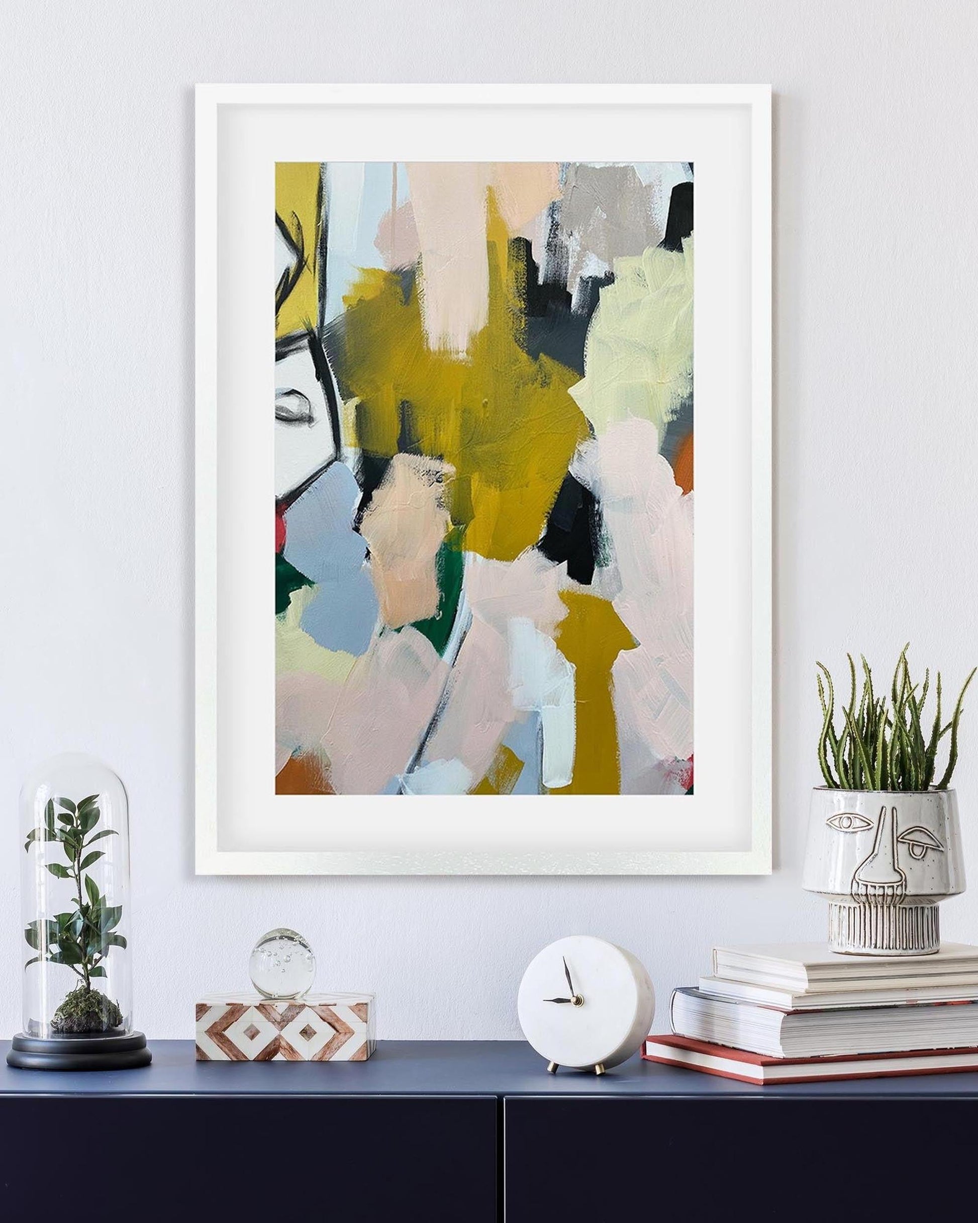 Abstract art print in a white frame on a wall above a shelf with decorative items.