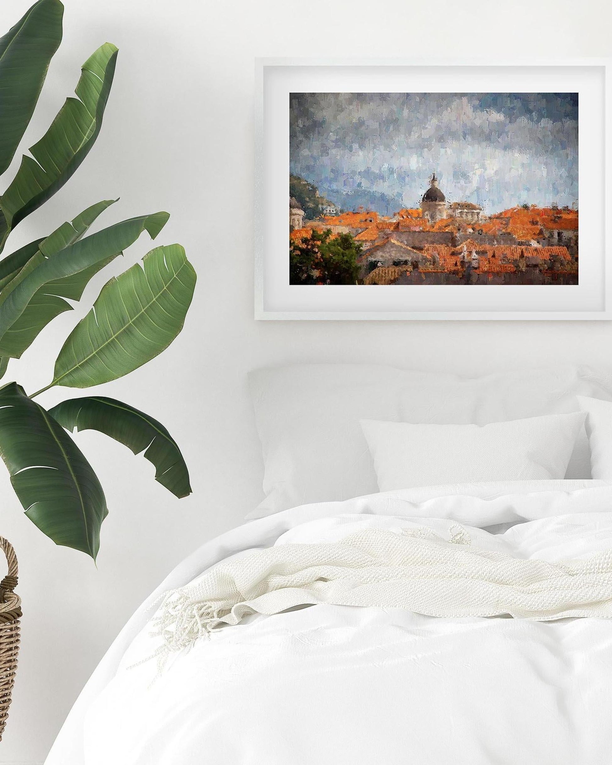 Framed artwork of a cityscape above a bed with a plant to the left.
