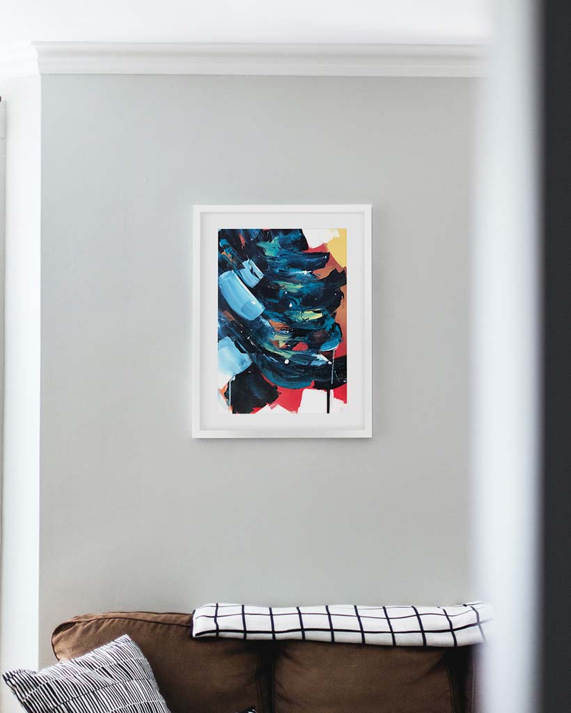 Abstract art print in a white frame on a light gray wall above a brown sofa.