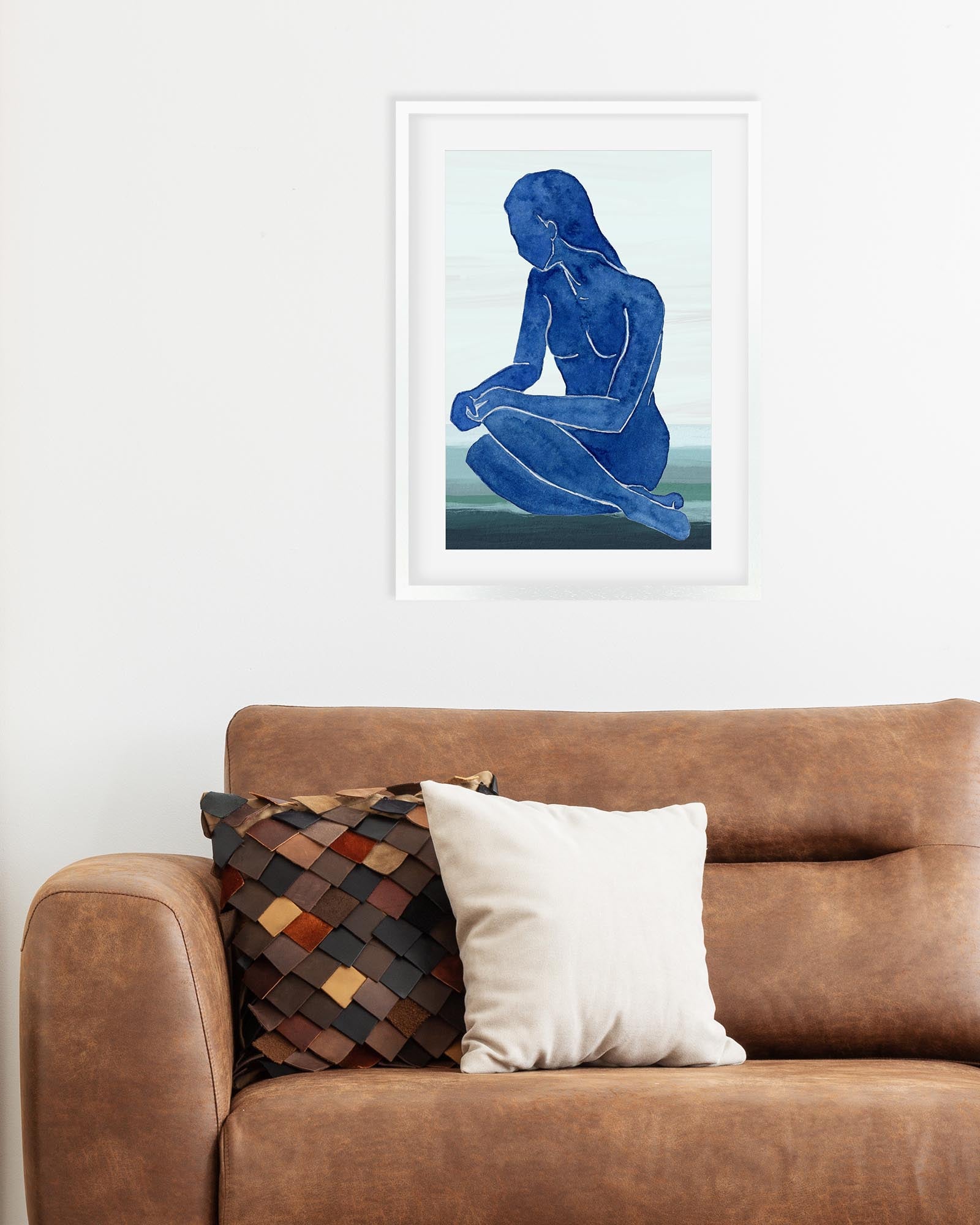 Framed artwork of a blue abstract figure on a white wall above a brown sofa with decorative pillows.