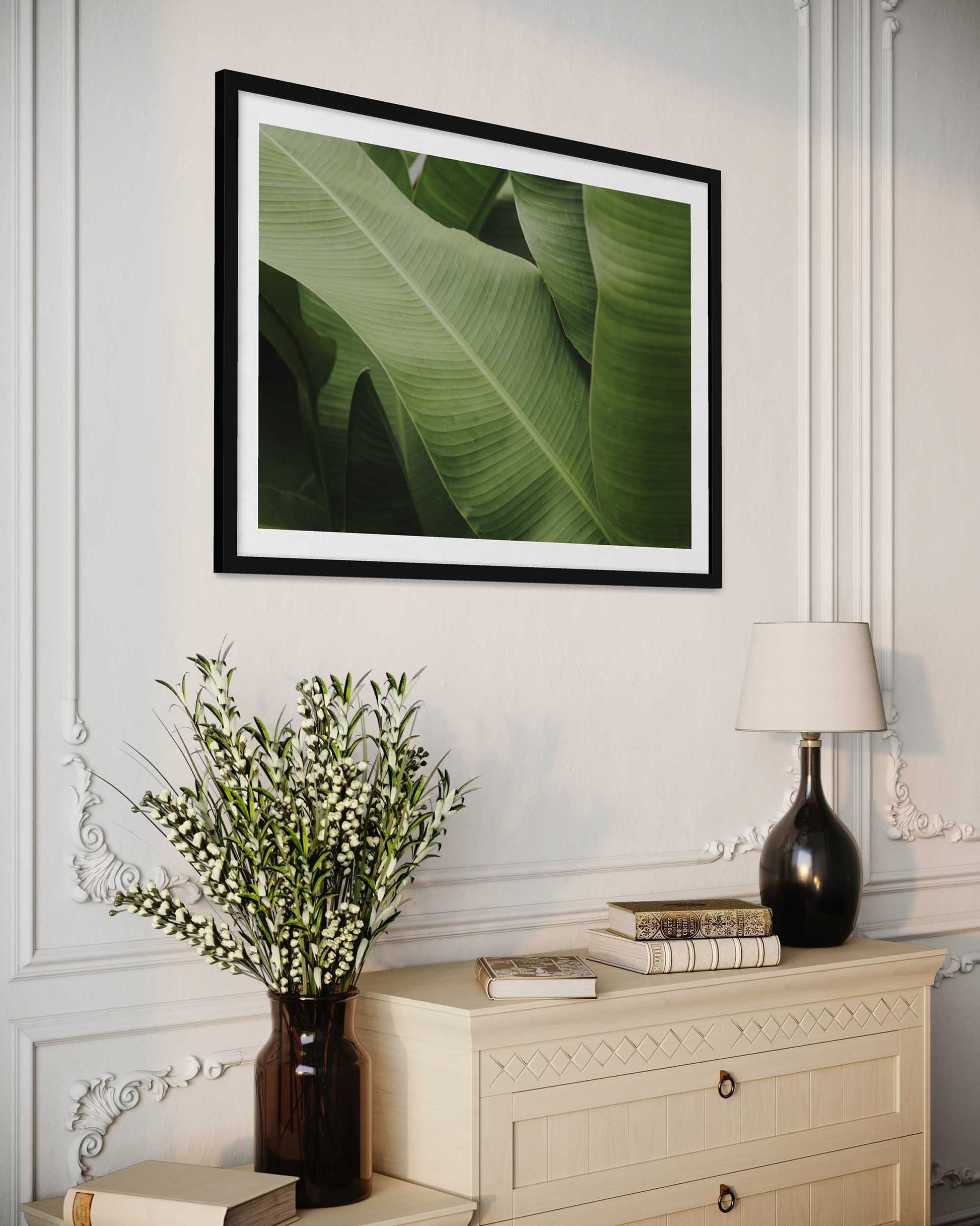 Framed green leaf print on a wall above a dresser with decorative items.