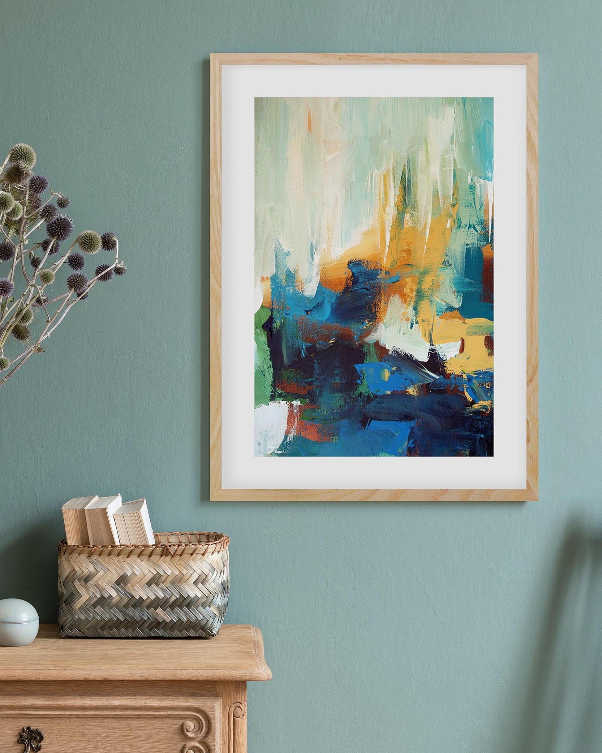 Framed abstract art on a wall with a vase of dried flowers and a basket below.