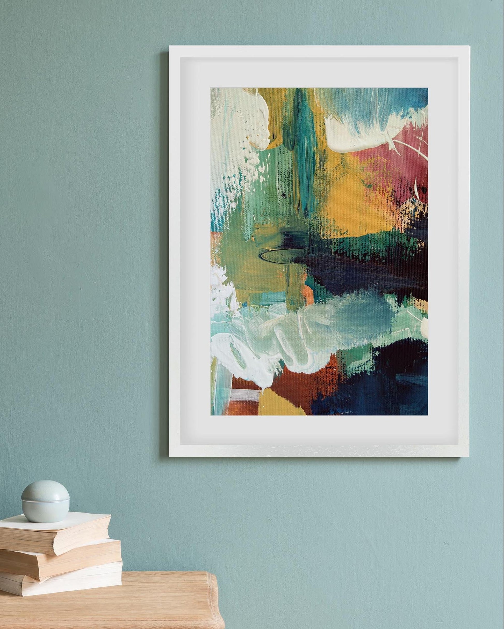 Framed abstract painting on a light blue wall with books and a plant below.
