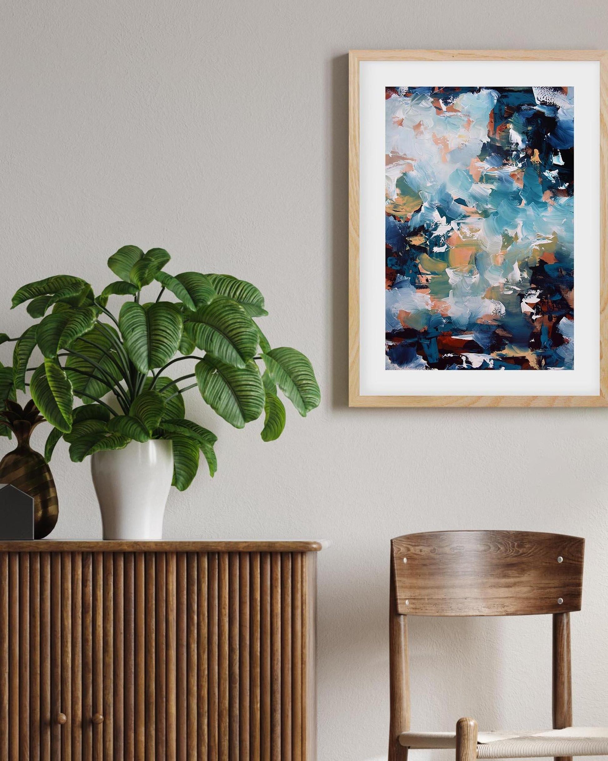 Framed abstract art on a wall with a plant and wooden furniture in the foreground.