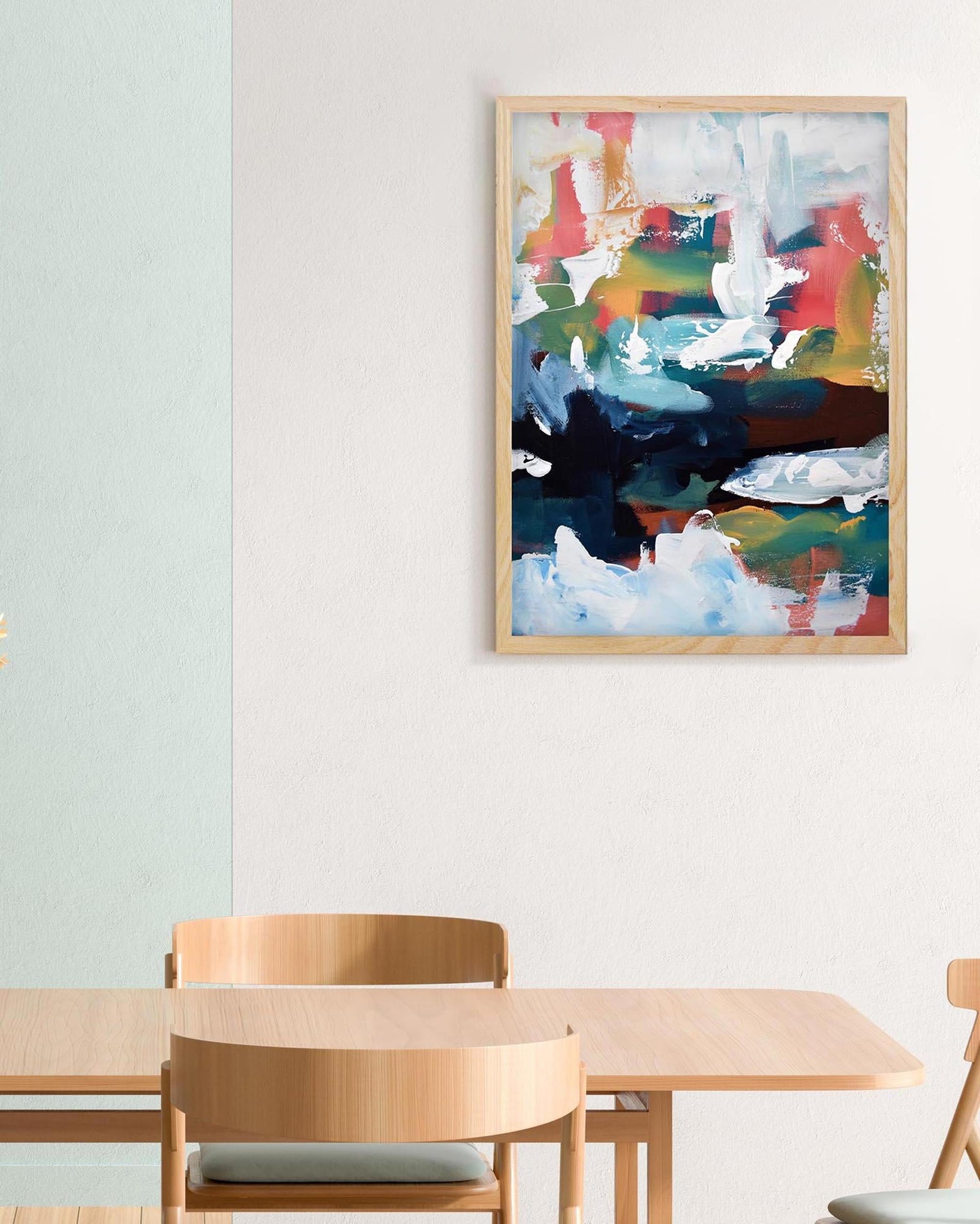 Abstract painting in a wooden frame above a dining table with chairs.