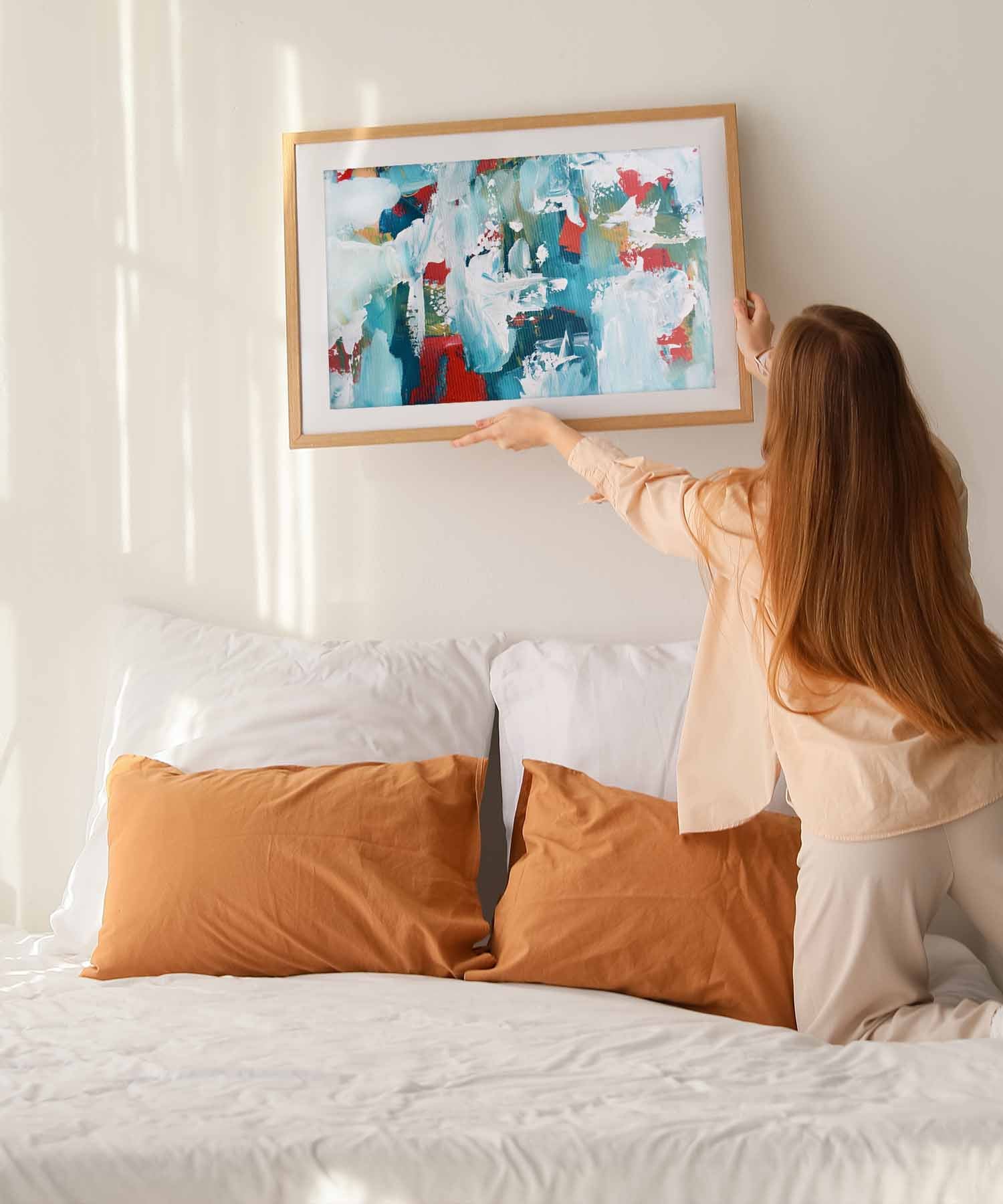 a woman hanging a colourful abstract wall art print above a bed