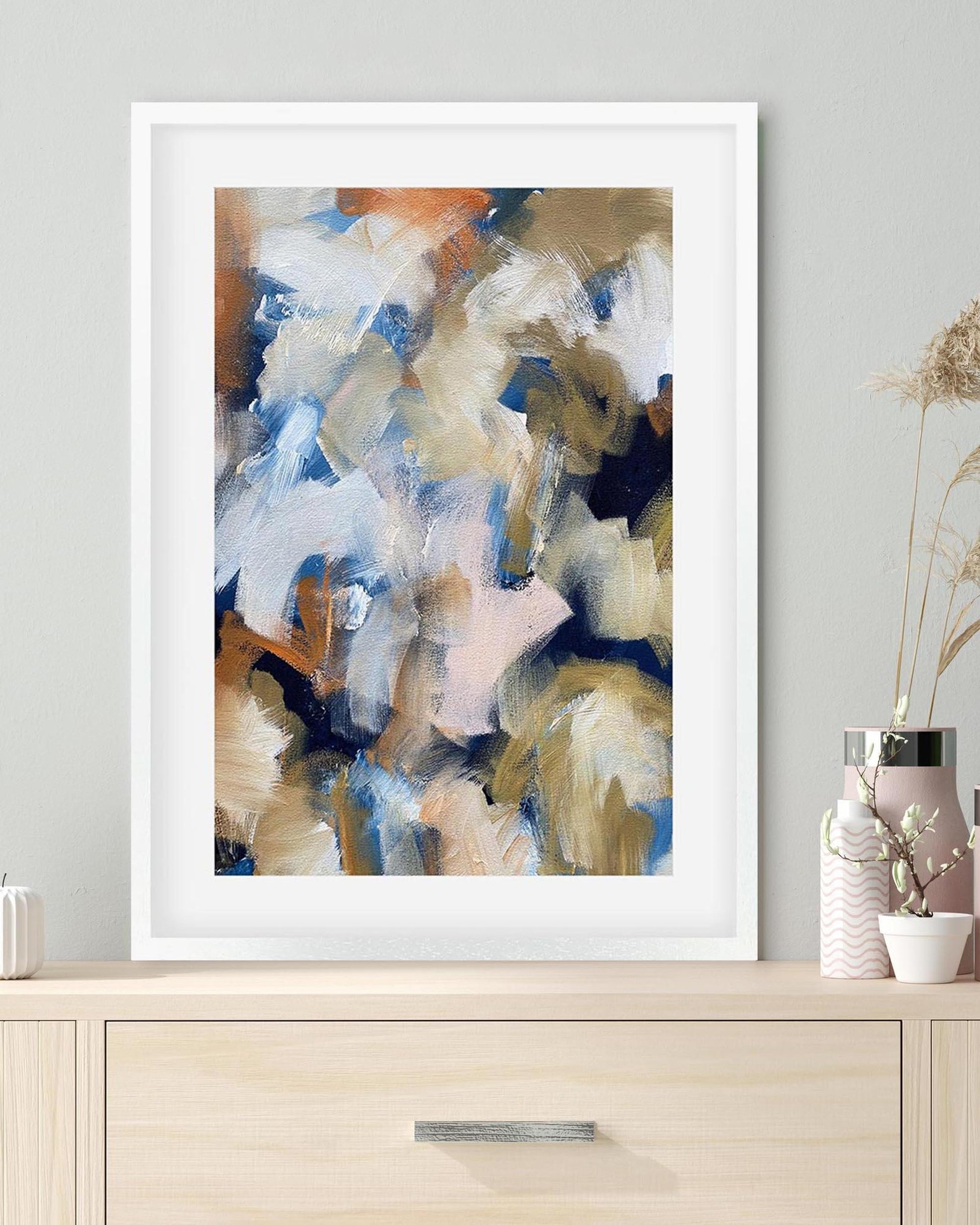 Framed abstract art print on a wall above a wooden cabinet.