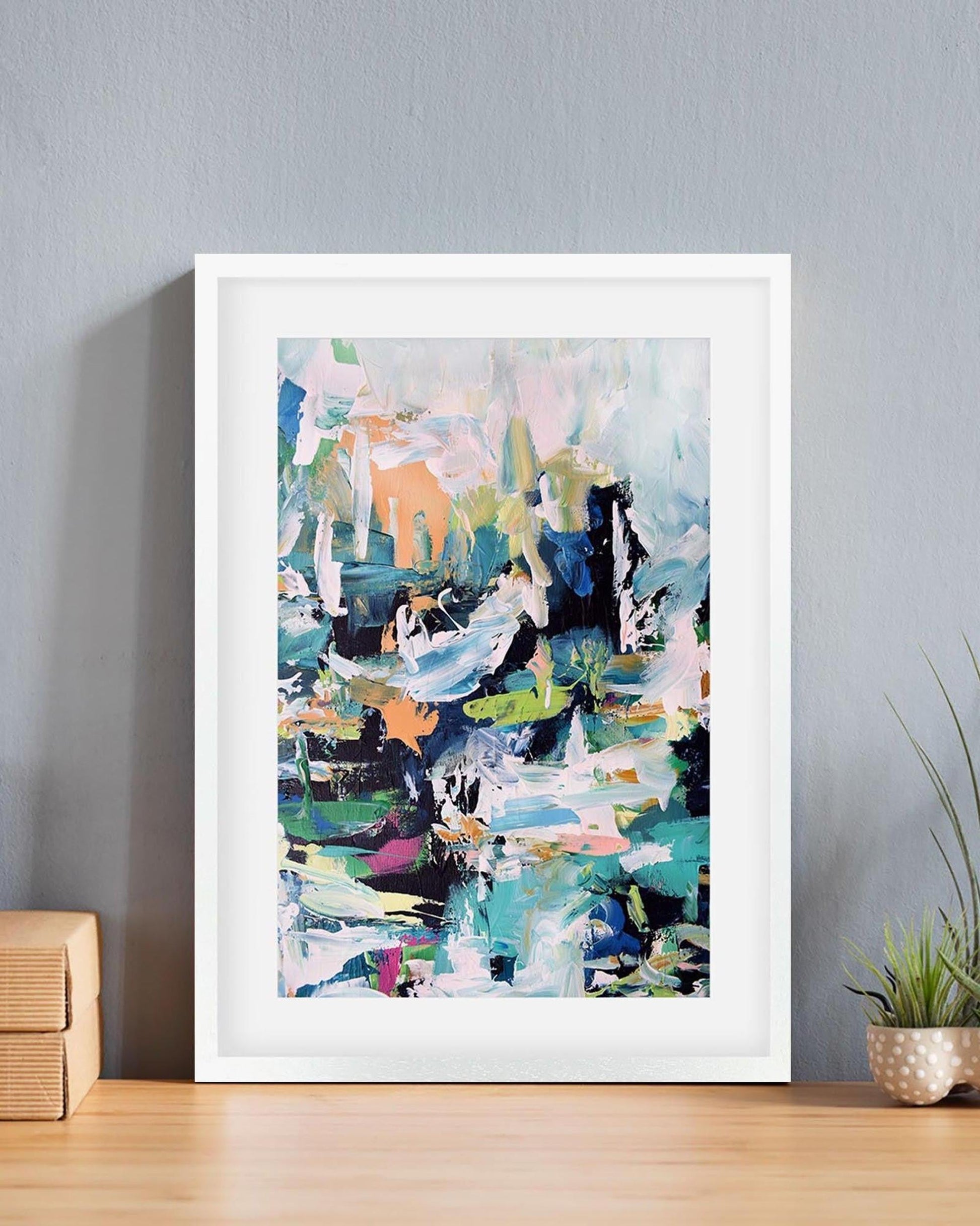 Framed abstract art print on a wall with a plant and wooden box below.