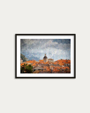 Framed painting of a cityscape with orange rooftops and a prominent building on a white wall.