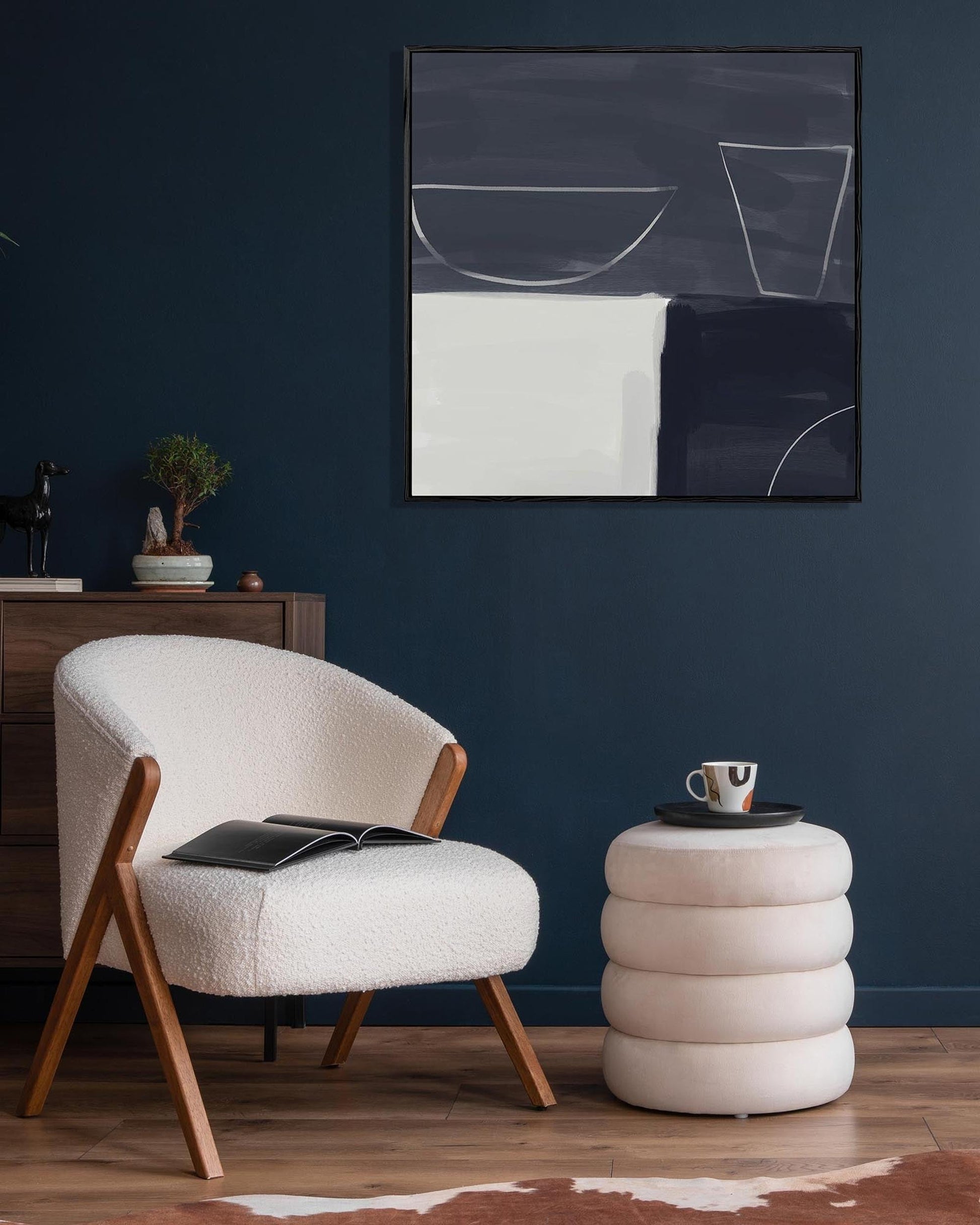 Modern interior with a white armchair, round side table, and abstract art on a dark blue wall.