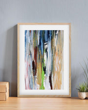 Framed abstract art print on a wall with a plant and wooden box below.