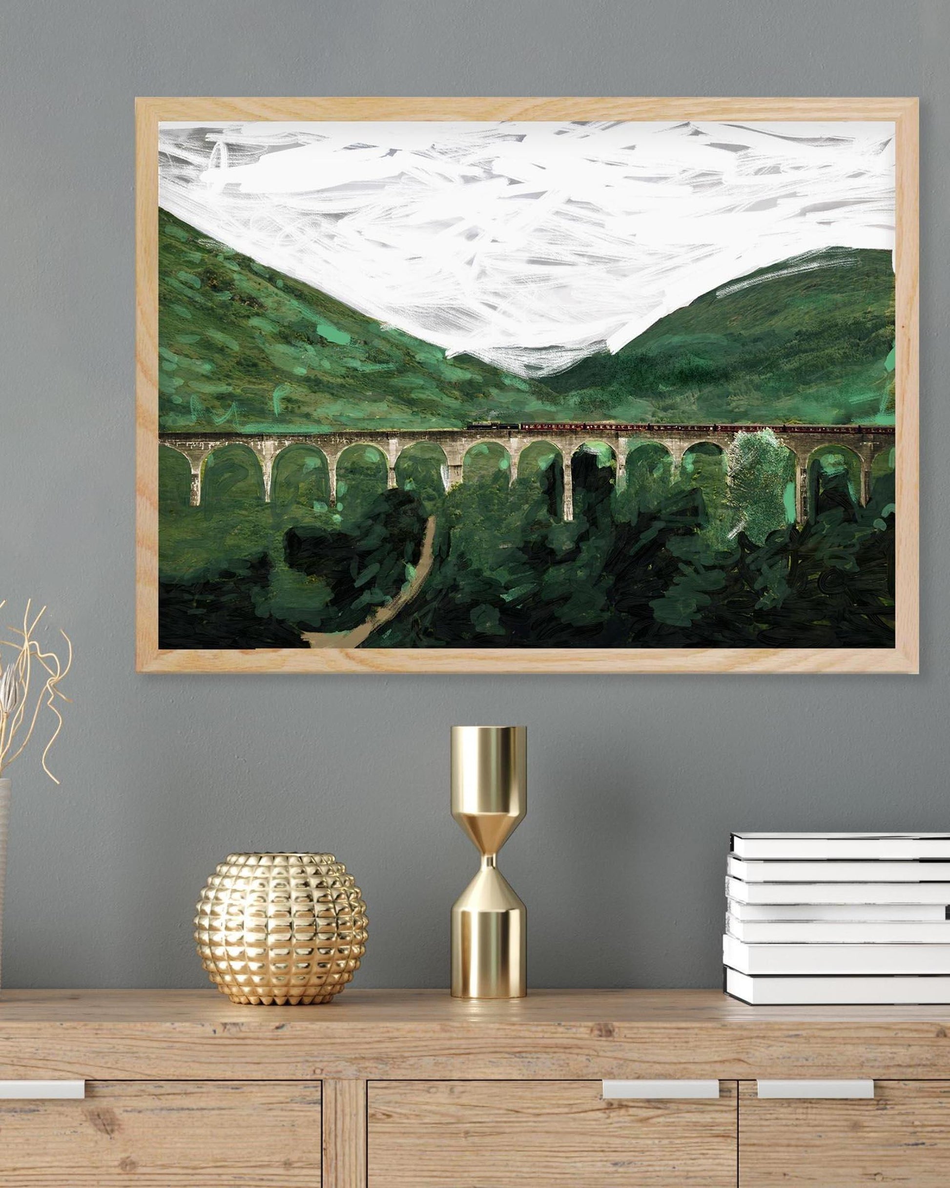 Framed landscape painting of a green valley with a bridge on a gray wall above a wooden cabinet.