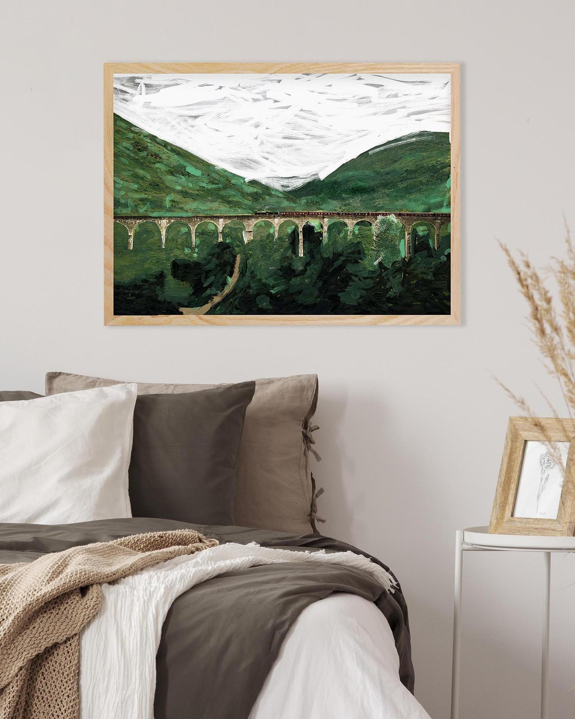 Framed artwork of a green landscape with a bridge on a wall above a bed.