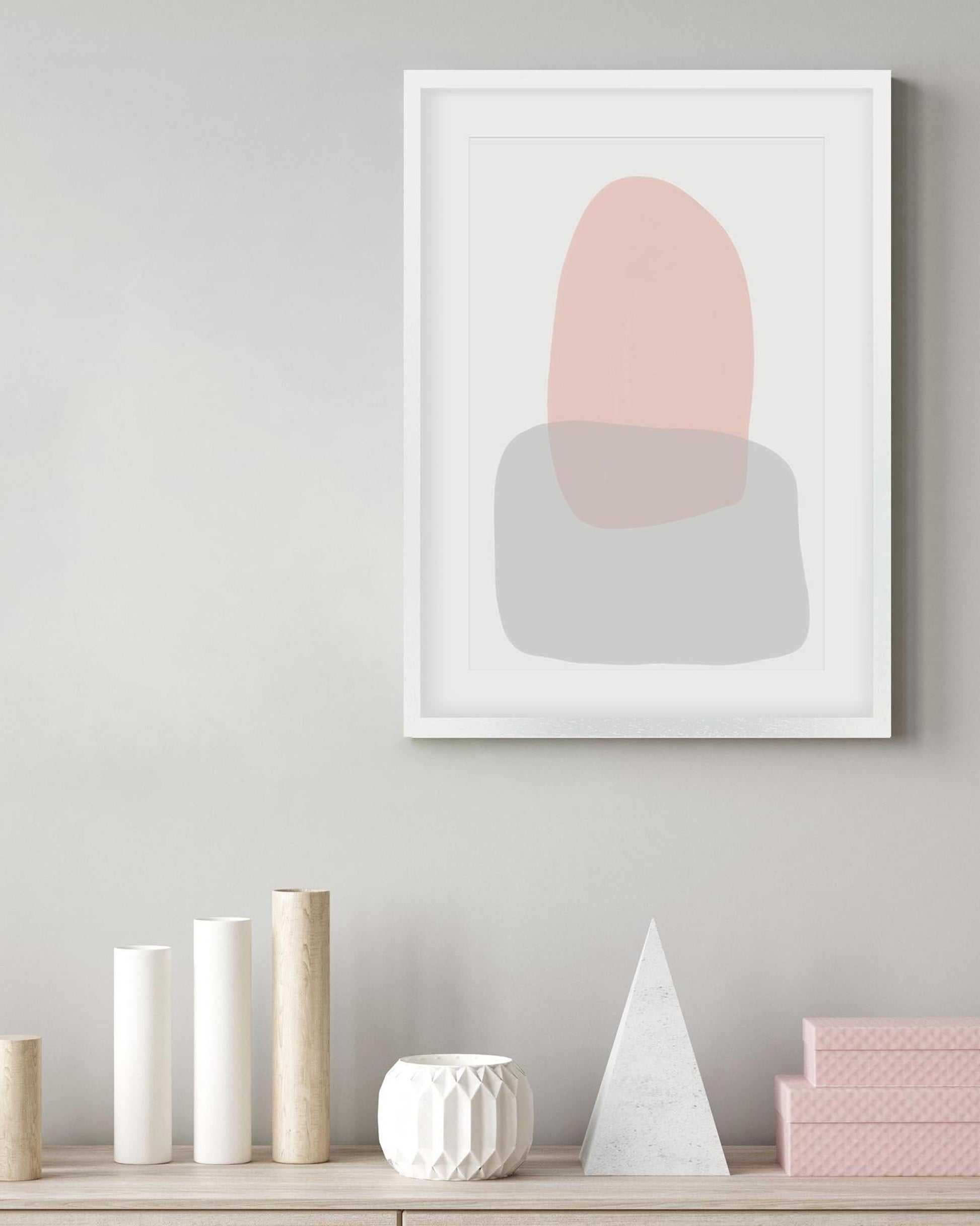 Framed abstract art print with pink and gray shapes on a wall above a shelf with decorative items.