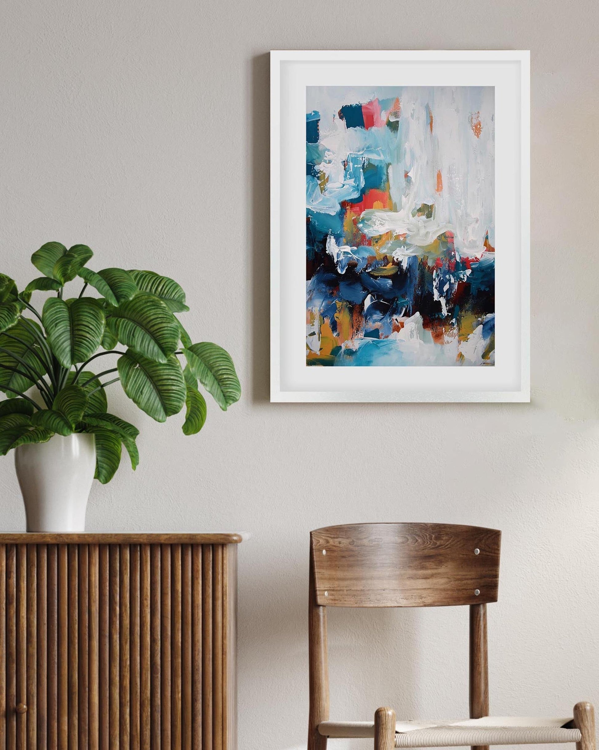 Framed abstract art piece on a wall with a plant and wooden furniture below.