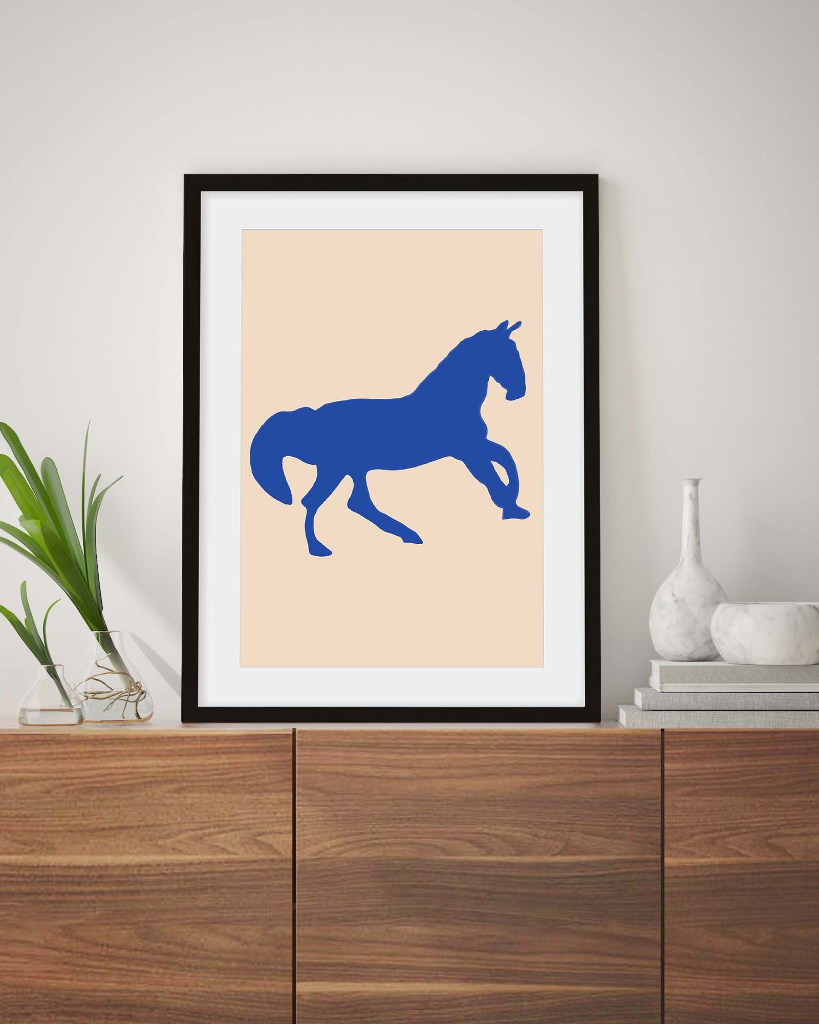 Framed poster with a blue horse silhouette on a beige background, displayed on a wooden cabinet.