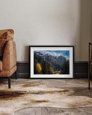 Framed landscape print on a wall in a room with a brown sofa and wooden floor.