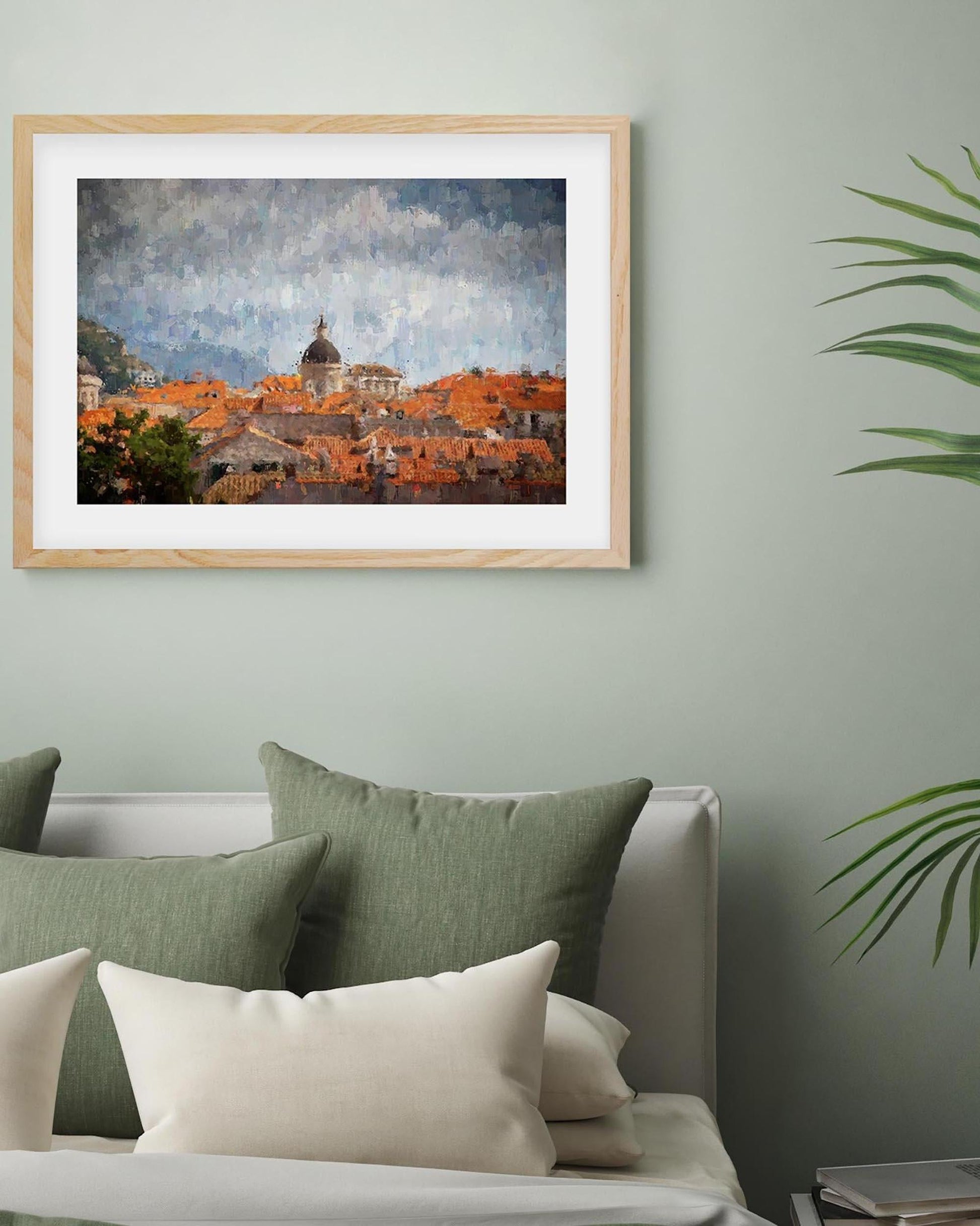 Framed artwork of a cityscape with orange buildings on a light green wall above a sofa with green and beige cushions.