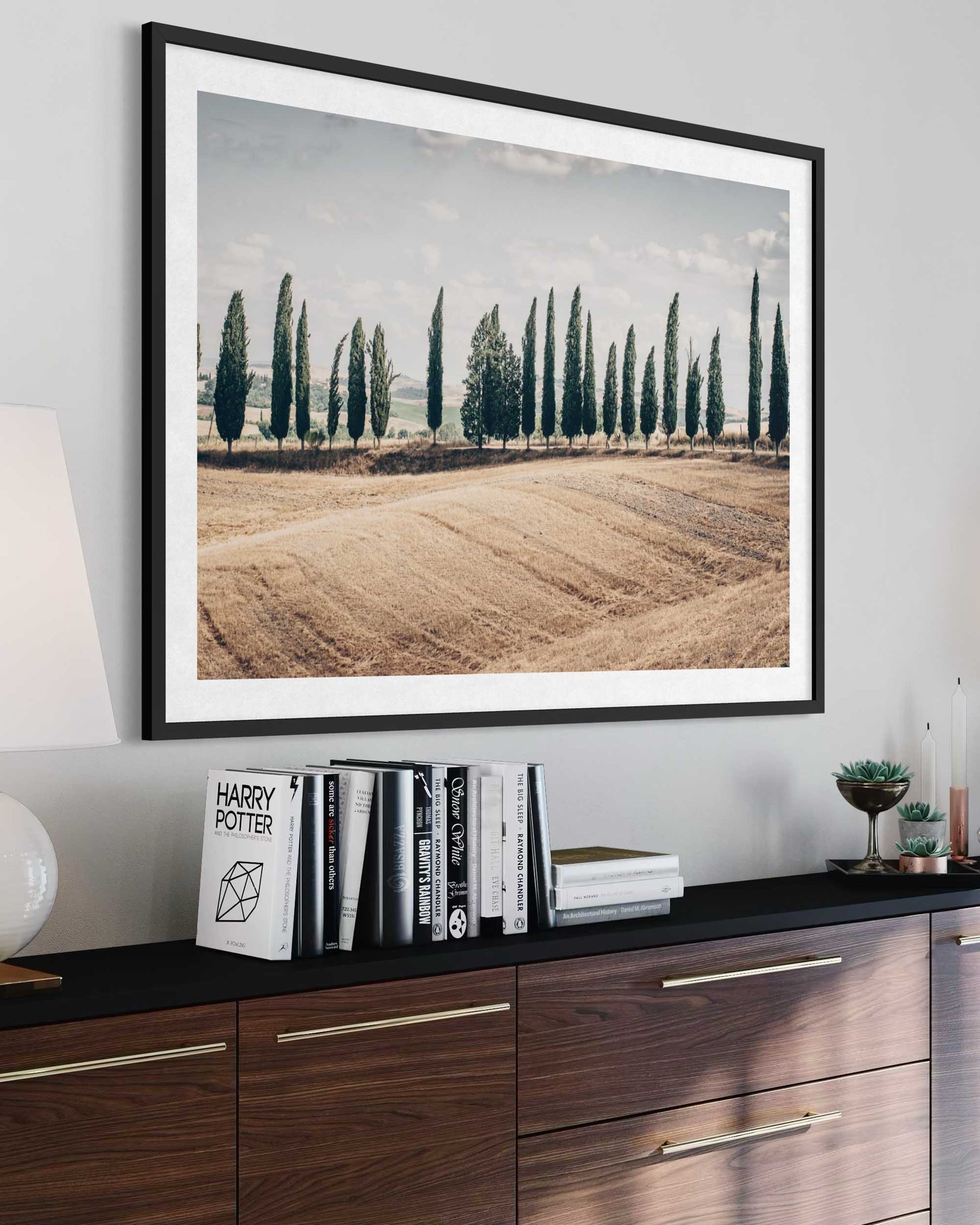 Framed landscape print of a field with trees on a wall above a wooden cabinet.