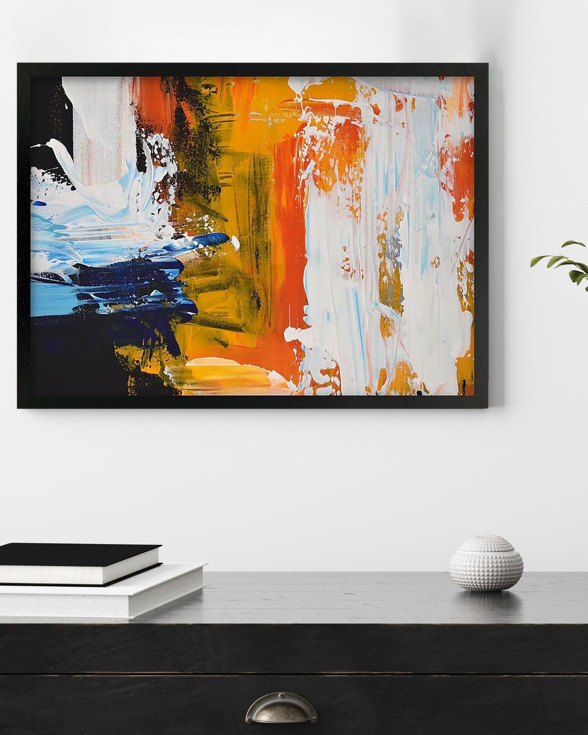 Abstract painting with orange, blue, and white on a black frame, displayed on a wall above a dark surface.
