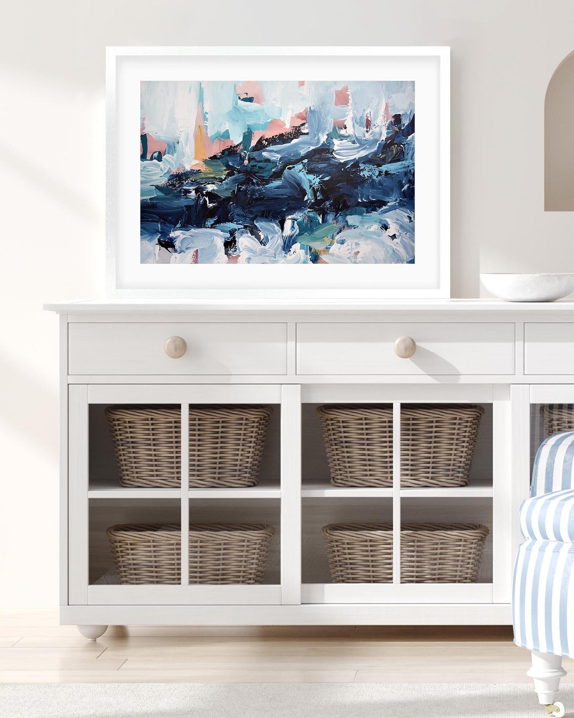 White cabinet with wicker baskets and a framed abstract painting in a room with a striped chair.