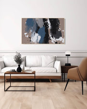 A large abstract framed canvas art hanging on a wall over a white sofa and a coffee table in a room with parquet flooring. The frame is beige, and the artwork features blocks of blue, black, and beige colors.
