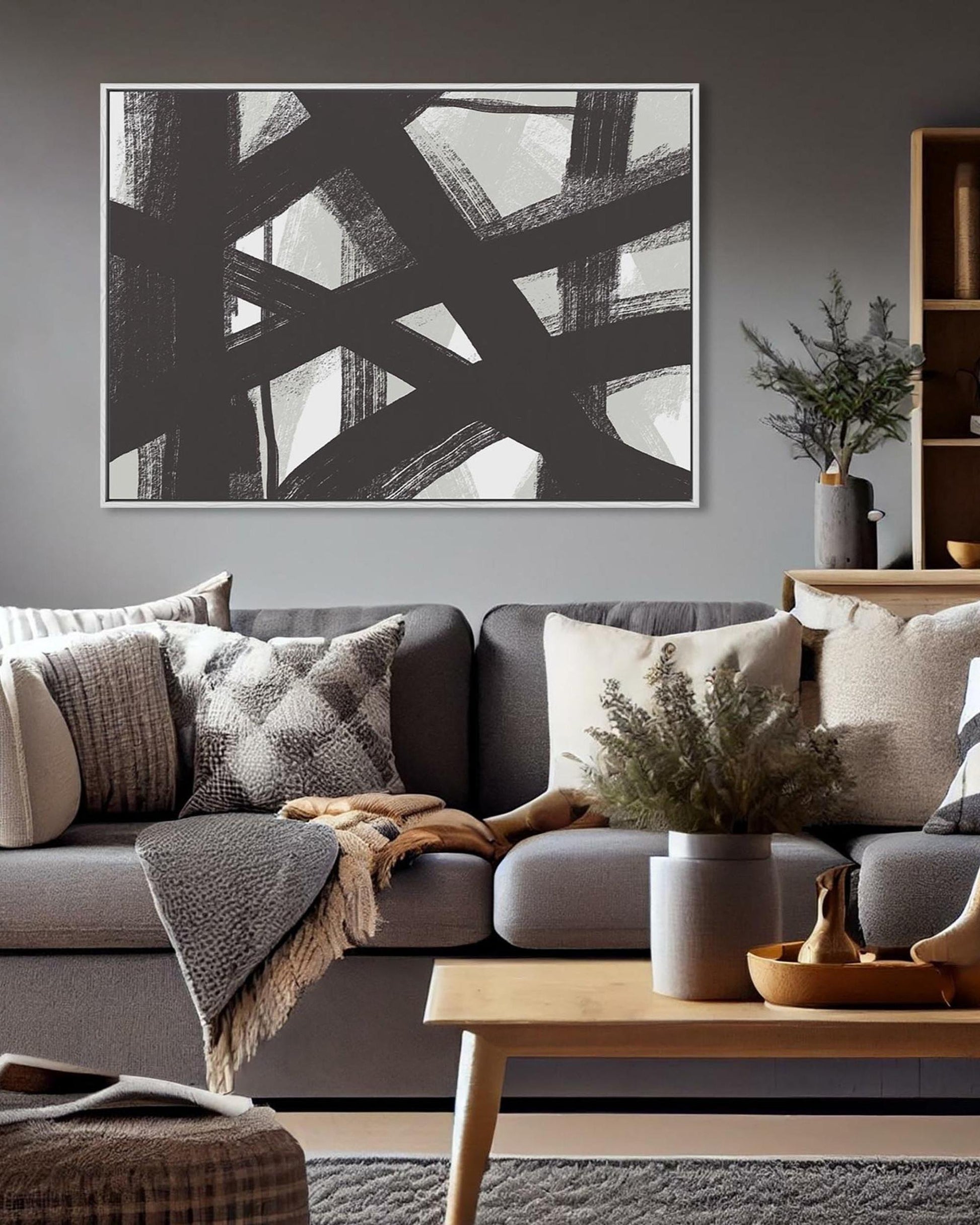 A framed canvas print with abstract black and white line marks hanging on a wall above a grey sofa with multiple pillows, in a living room setting.