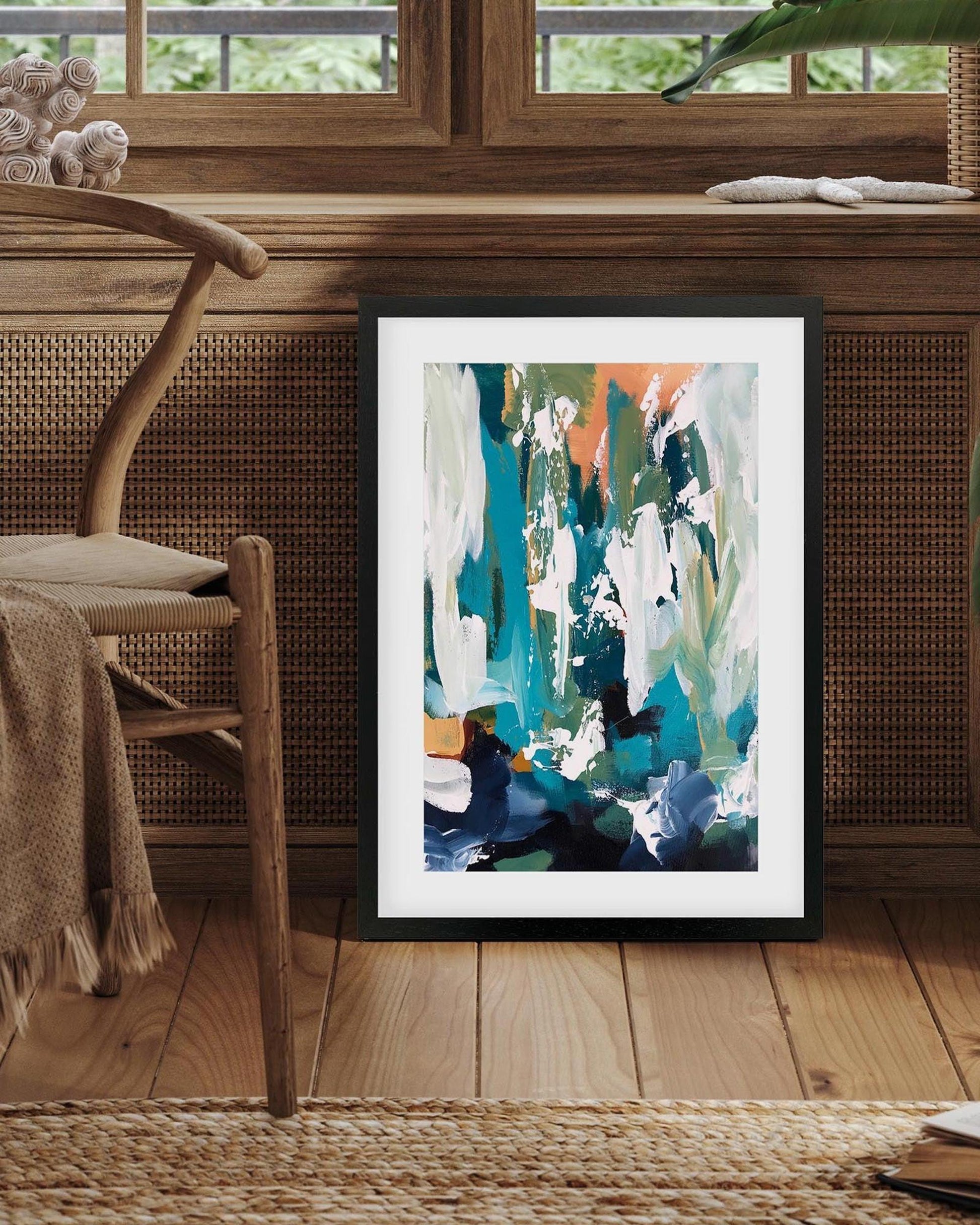 Framed abstract art piece on a wooden floor with a chair and wicker basket in the background.