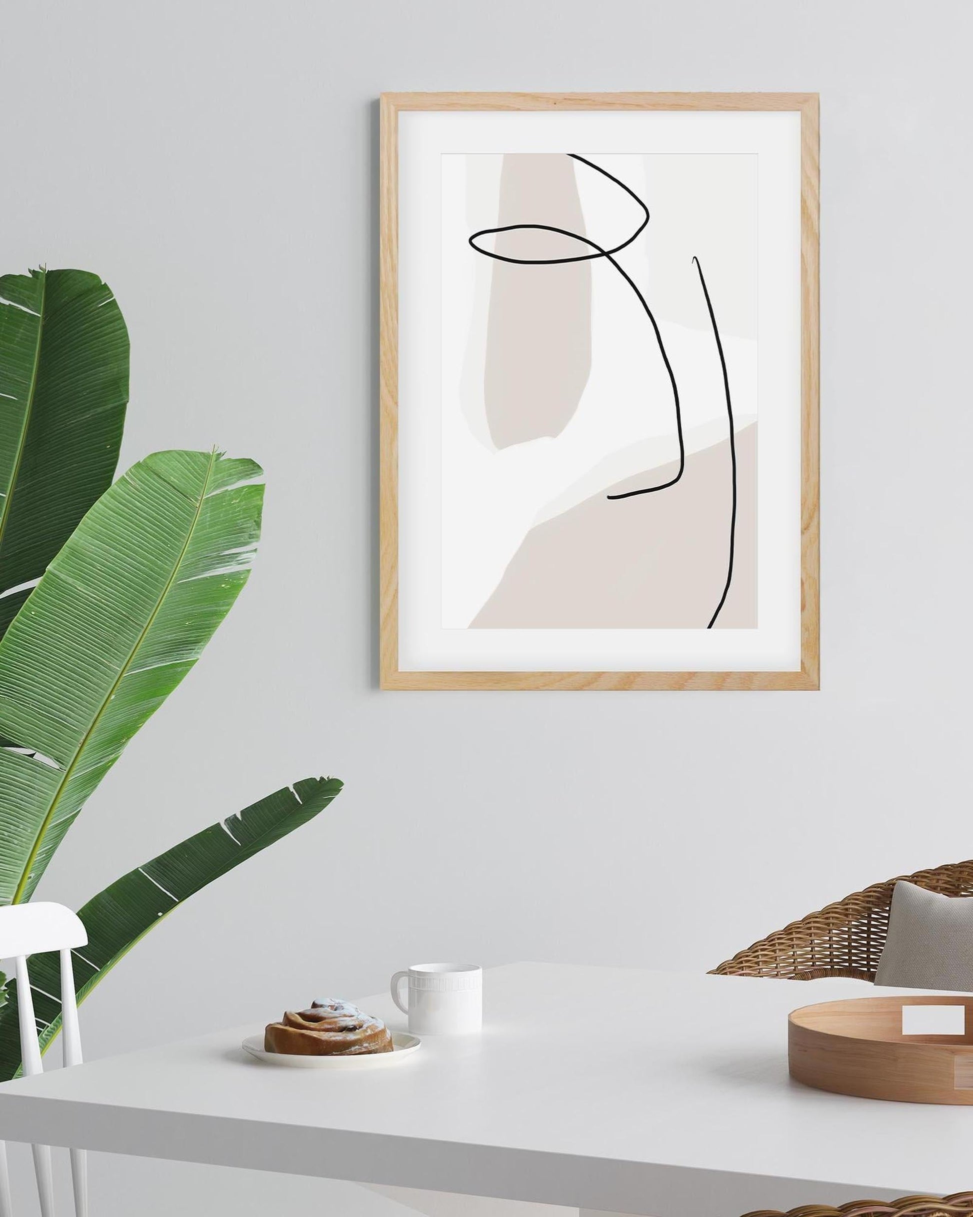 Abstract art print in a wooden frame on a wall with a plant and table in the foreground.