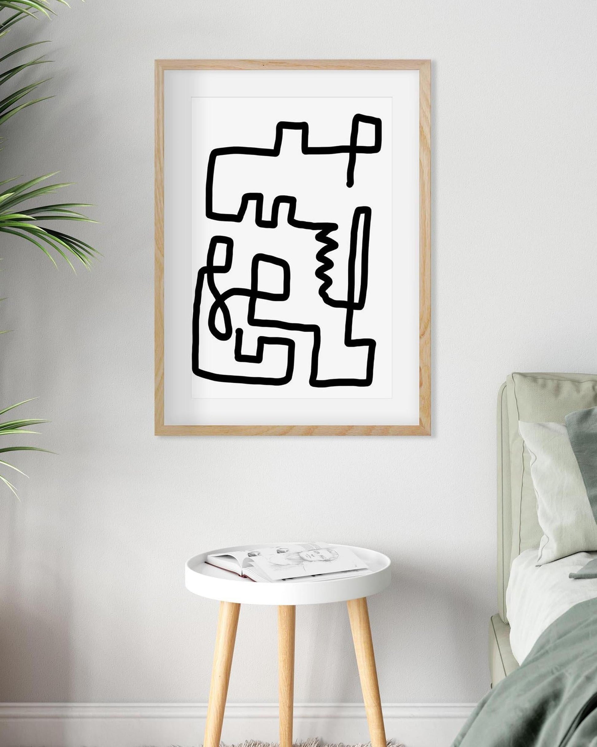 Abstract black line art in a wooden frame on a white wall above a stool.