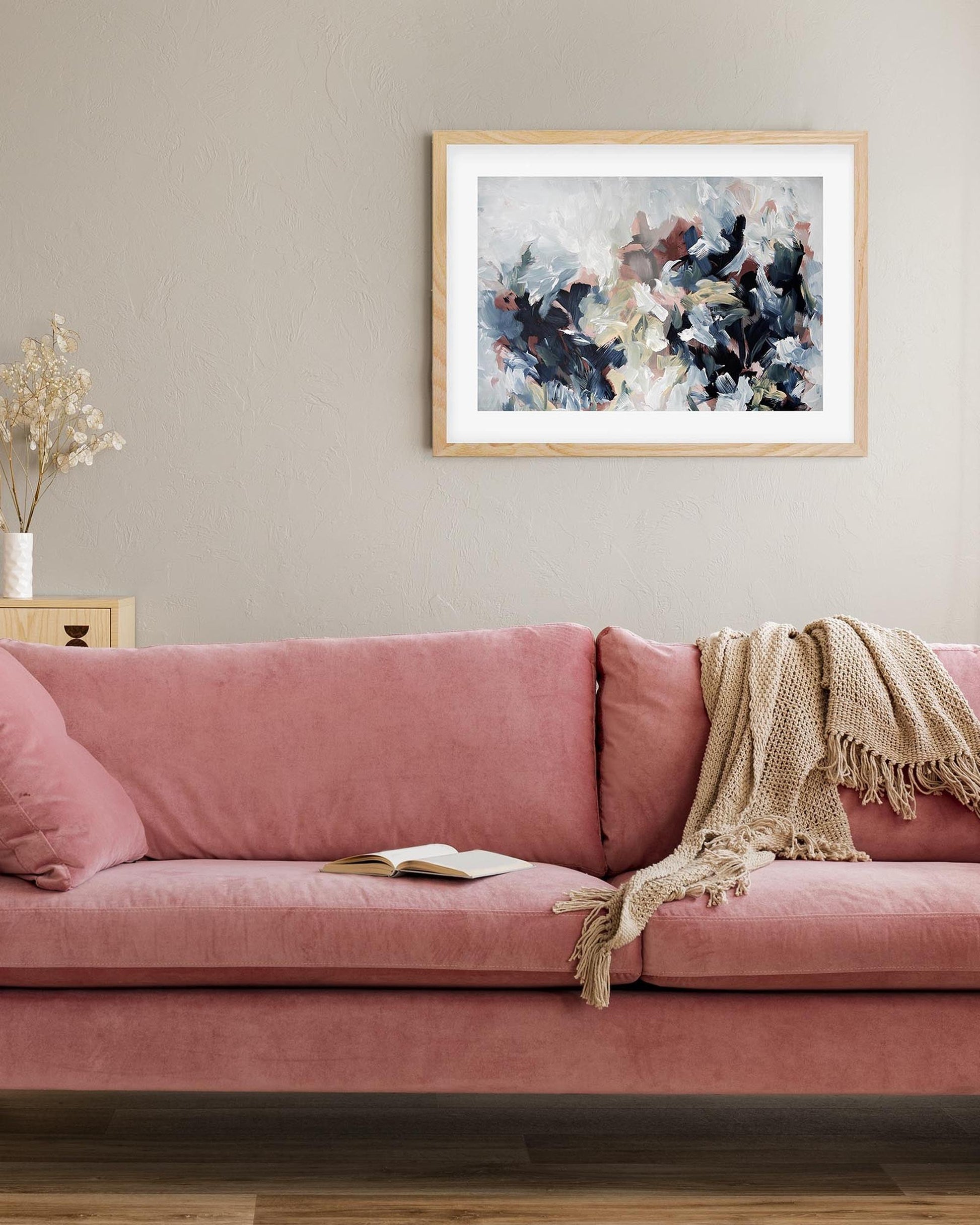 Pink sofa with a blanket and a framed abstract painting on a beige wall.