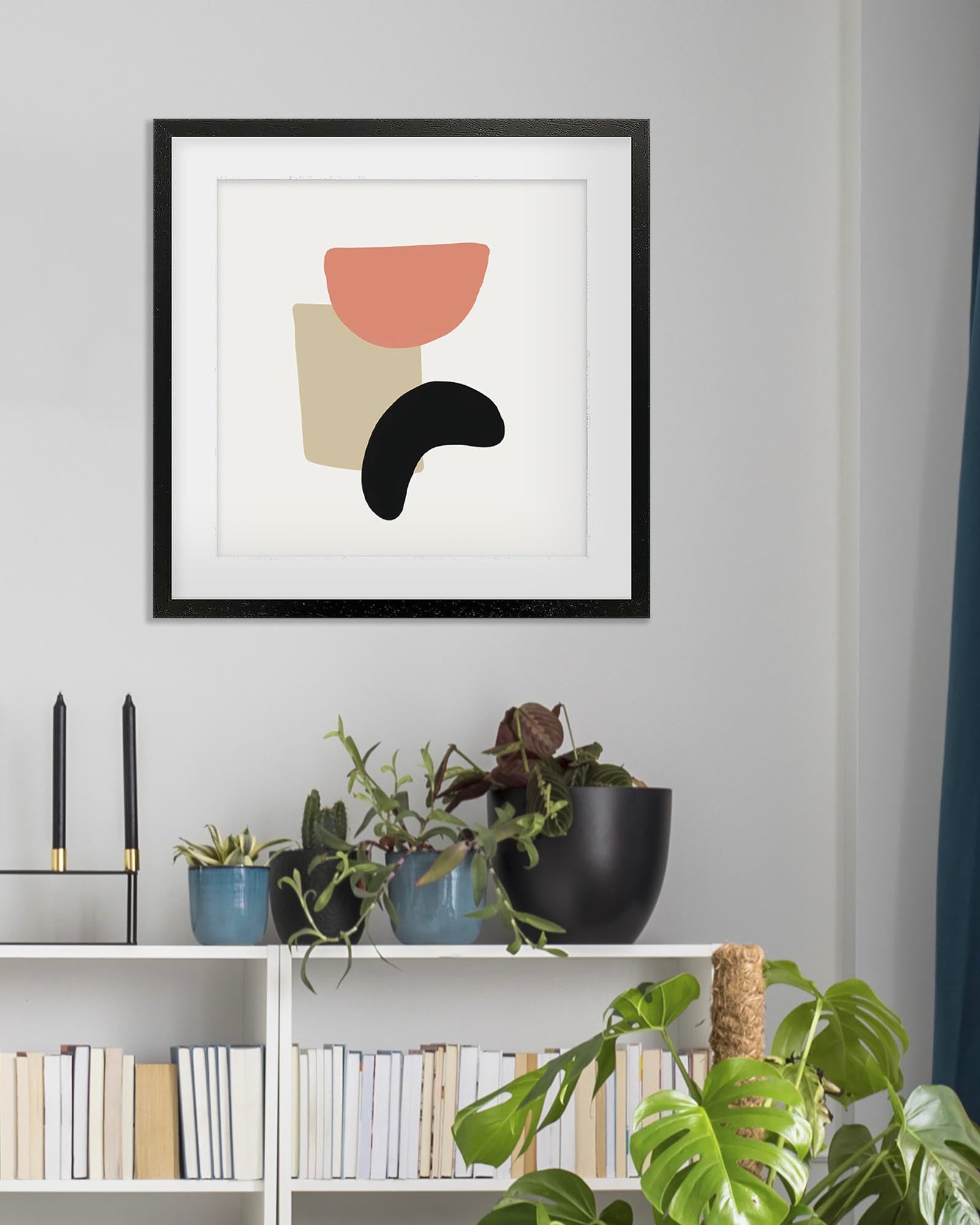 Framed abstract art print on a wall above a shelf with books and plants.