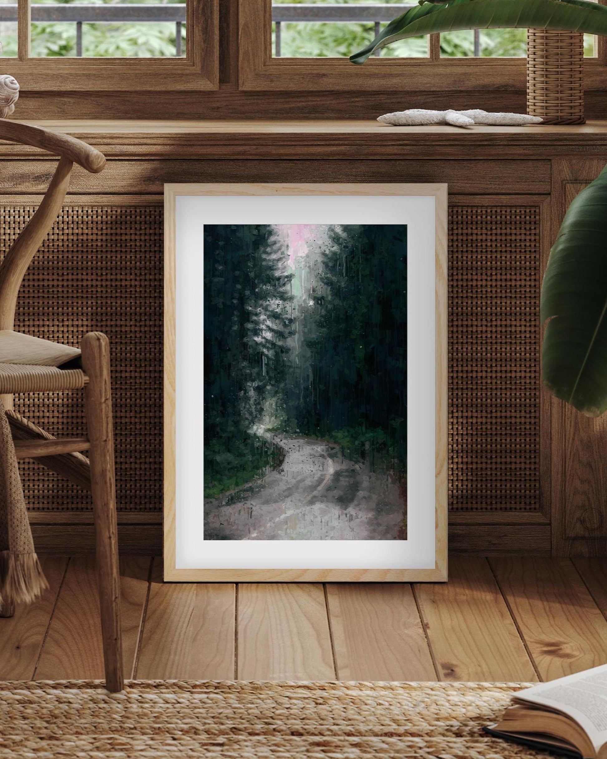Framed print of a forest path on a wooden floor with a chair and plant in the background.