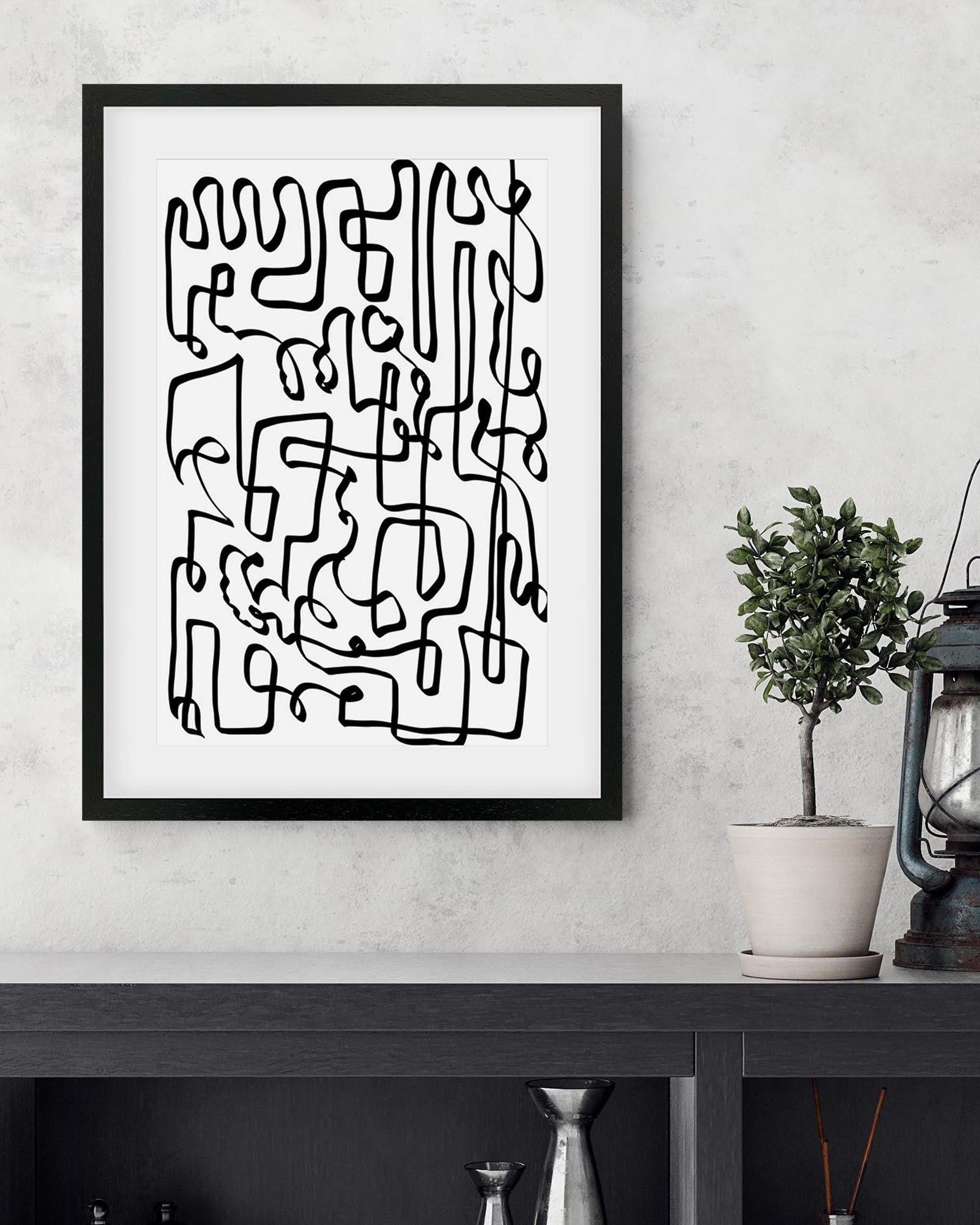 Framed abstract black line art on a white wall with a plant and lantern in the background.
