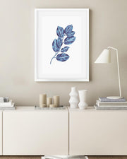 Framed artwork of blue leaves on a beige wall above a white cabinet with decorative items.
