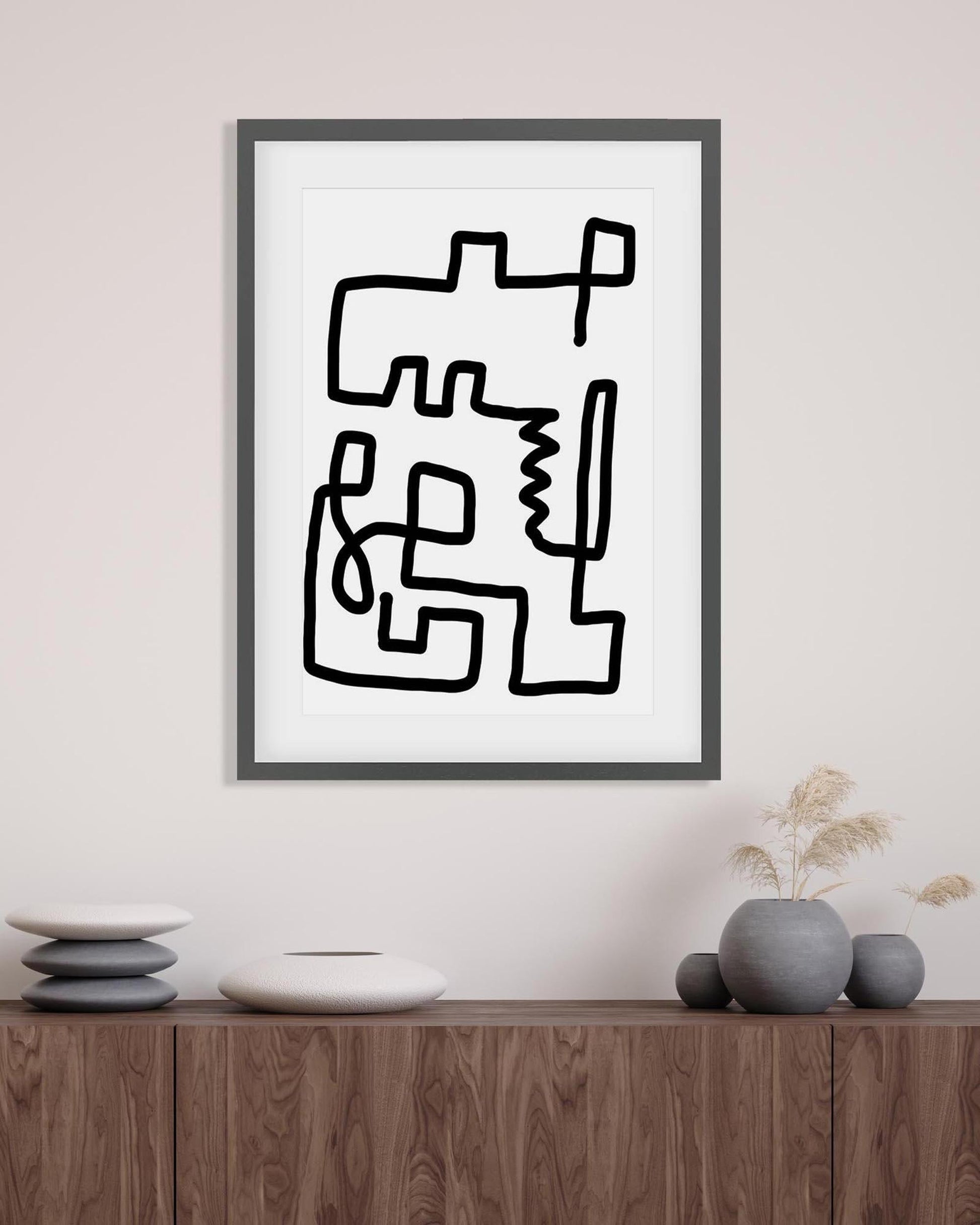 Framed abstract black line art on a white background, displayed on a wall above a wooden cabinet.