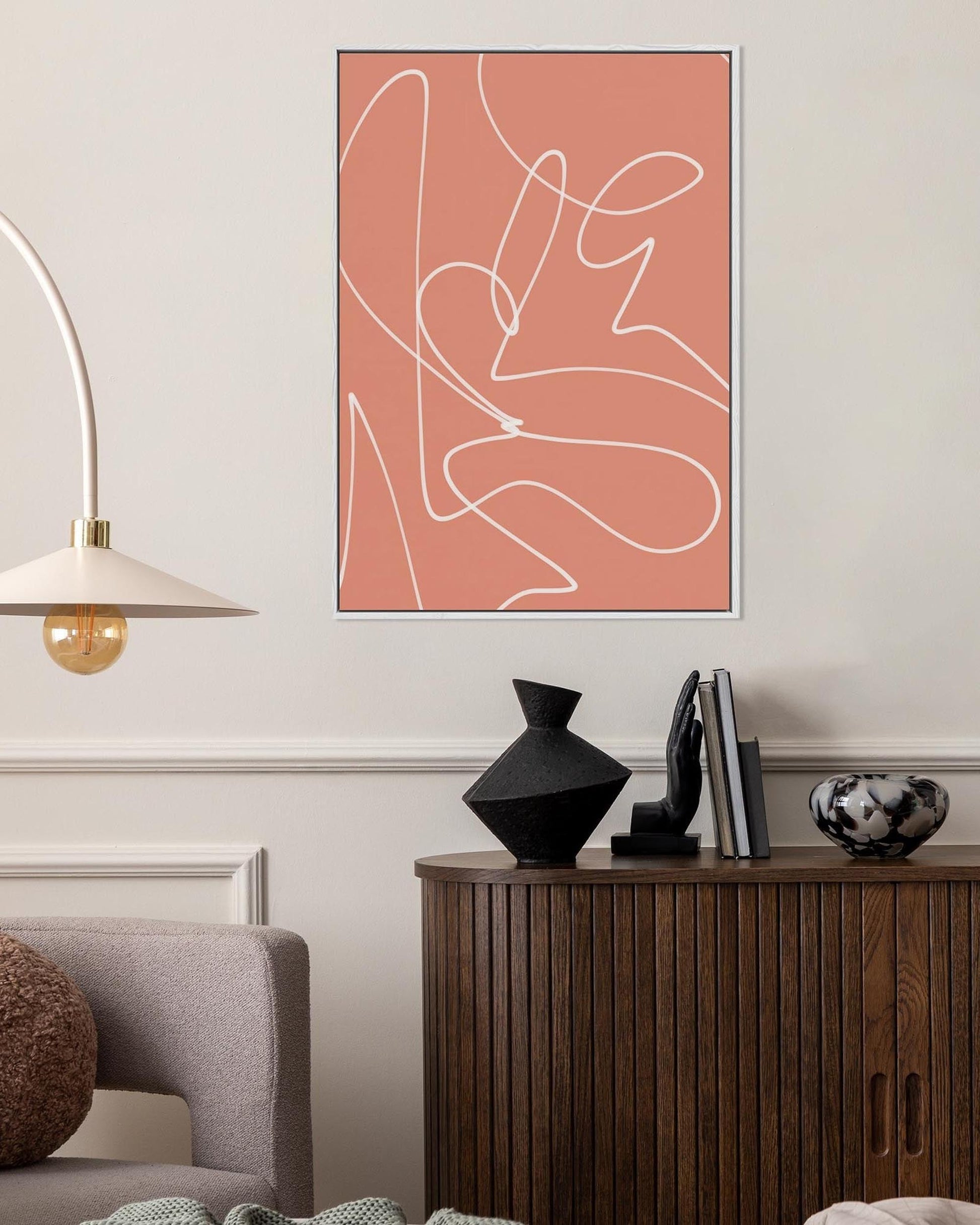 An abstract curved lines framed canvas art hanging on a wall above a wooden sideboard in a room with various decor.
