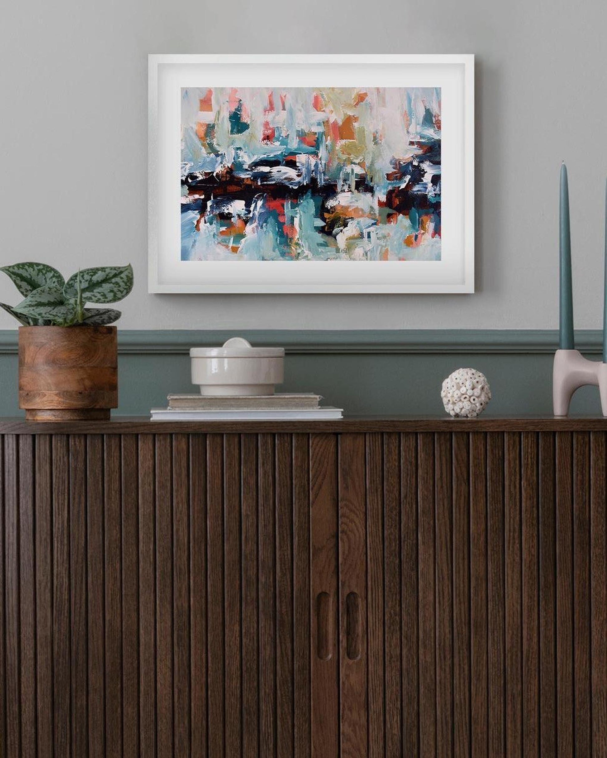 Framed abstract painting on a wall above a wooden cabinet with decorative items.