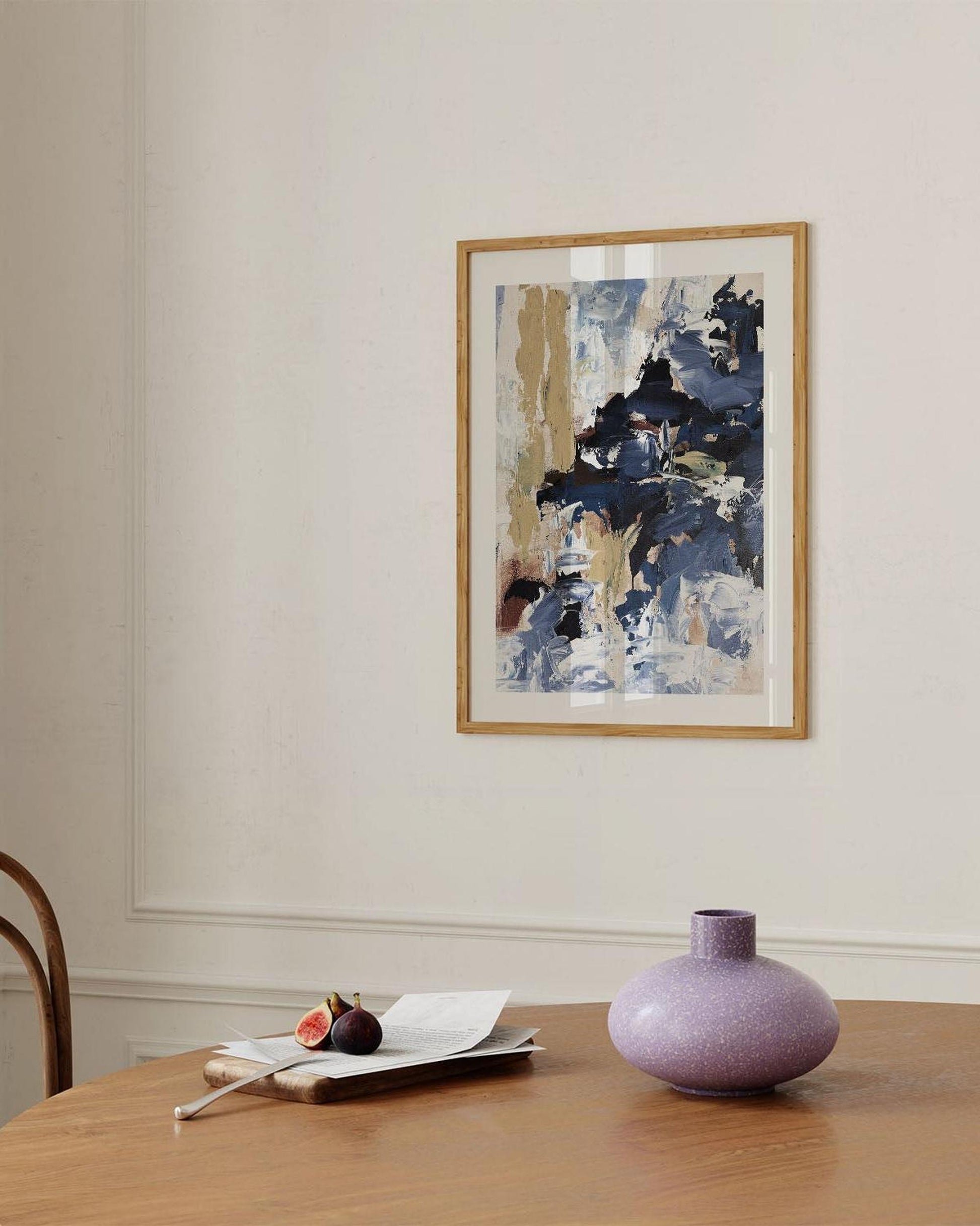 Abstract painting on a wall above a table with a vase and fruit.