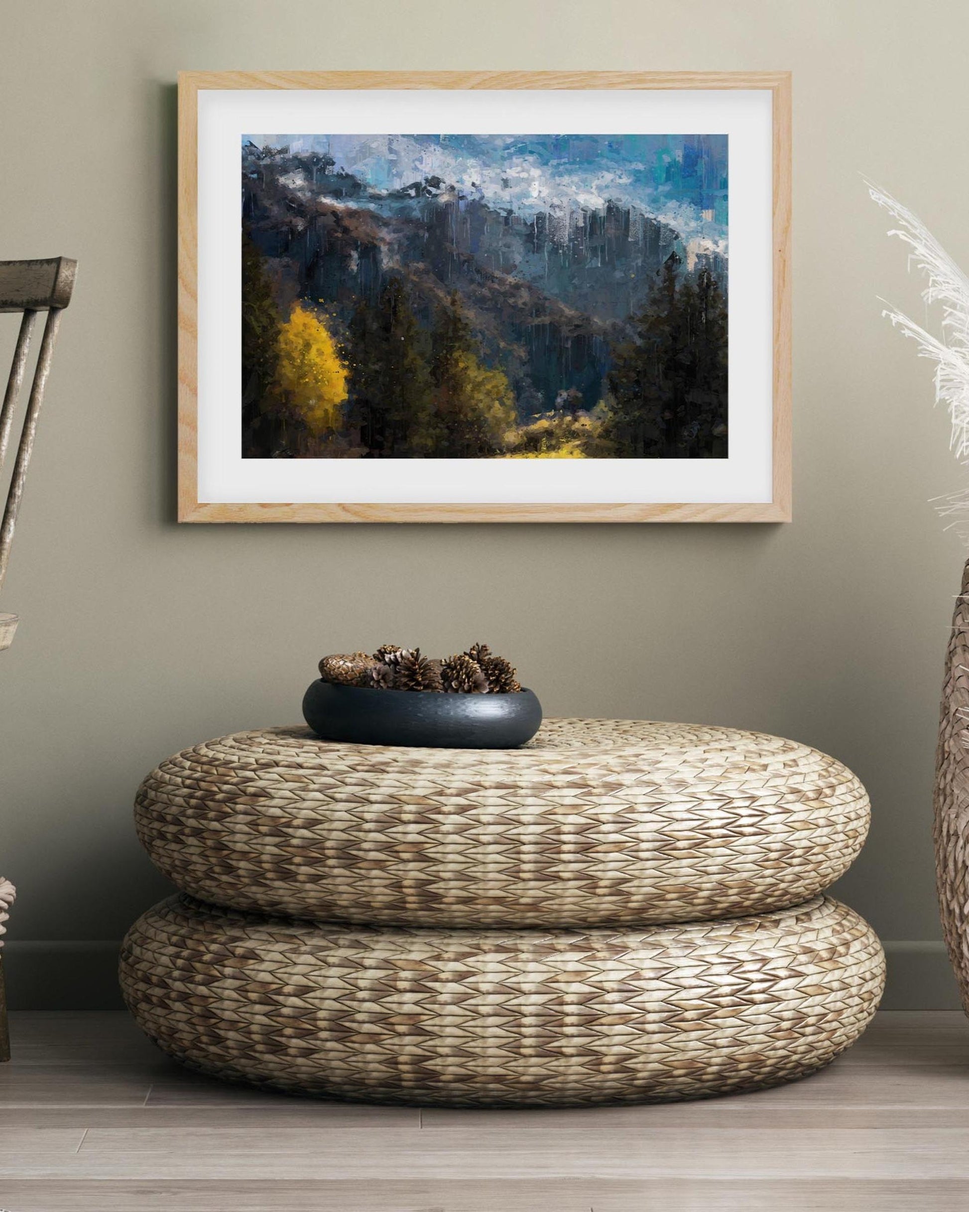Framed landscape painting above a woven ottoman in a room.
