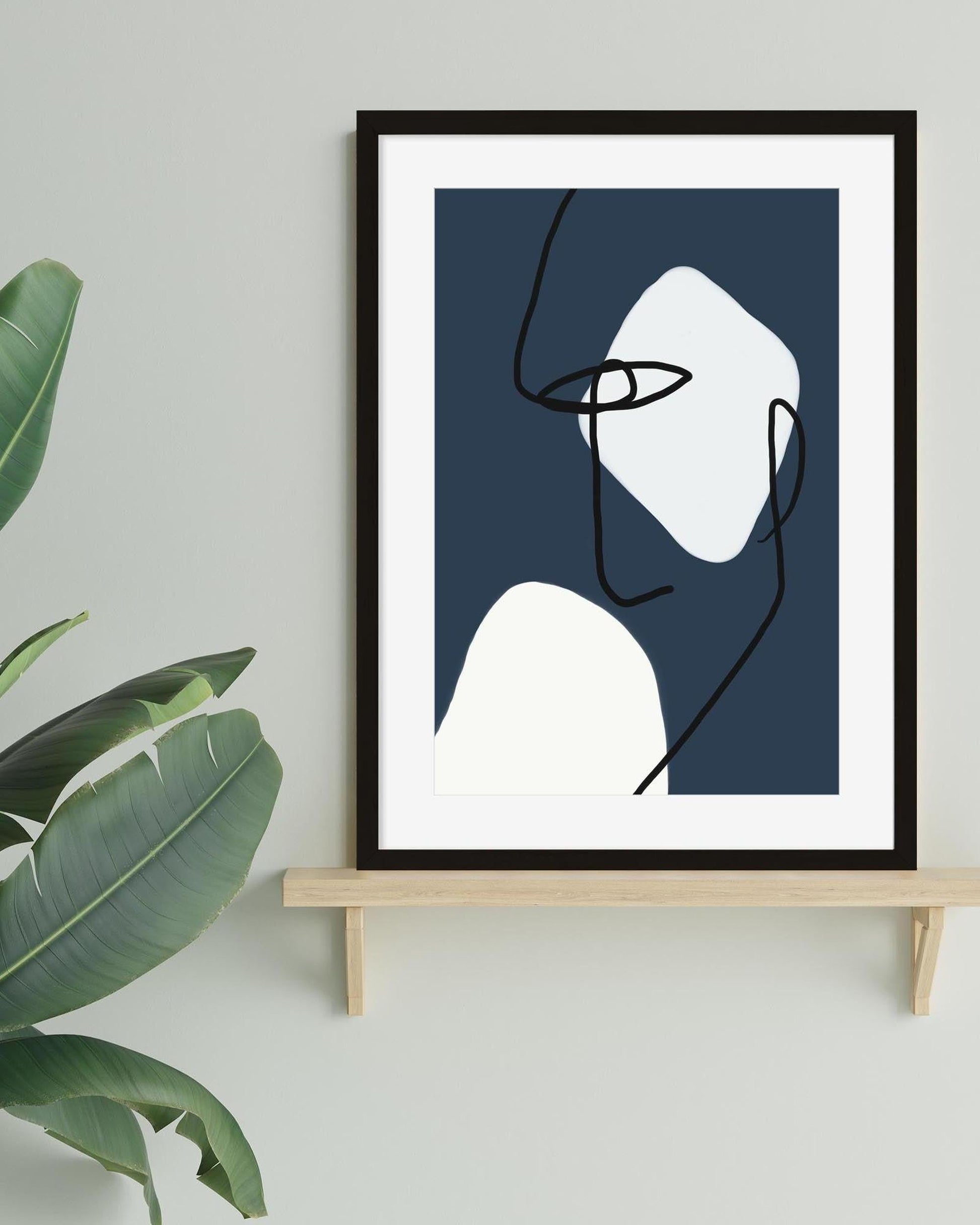 Framed abstract art print of a face on a white wall with a plant to the left.