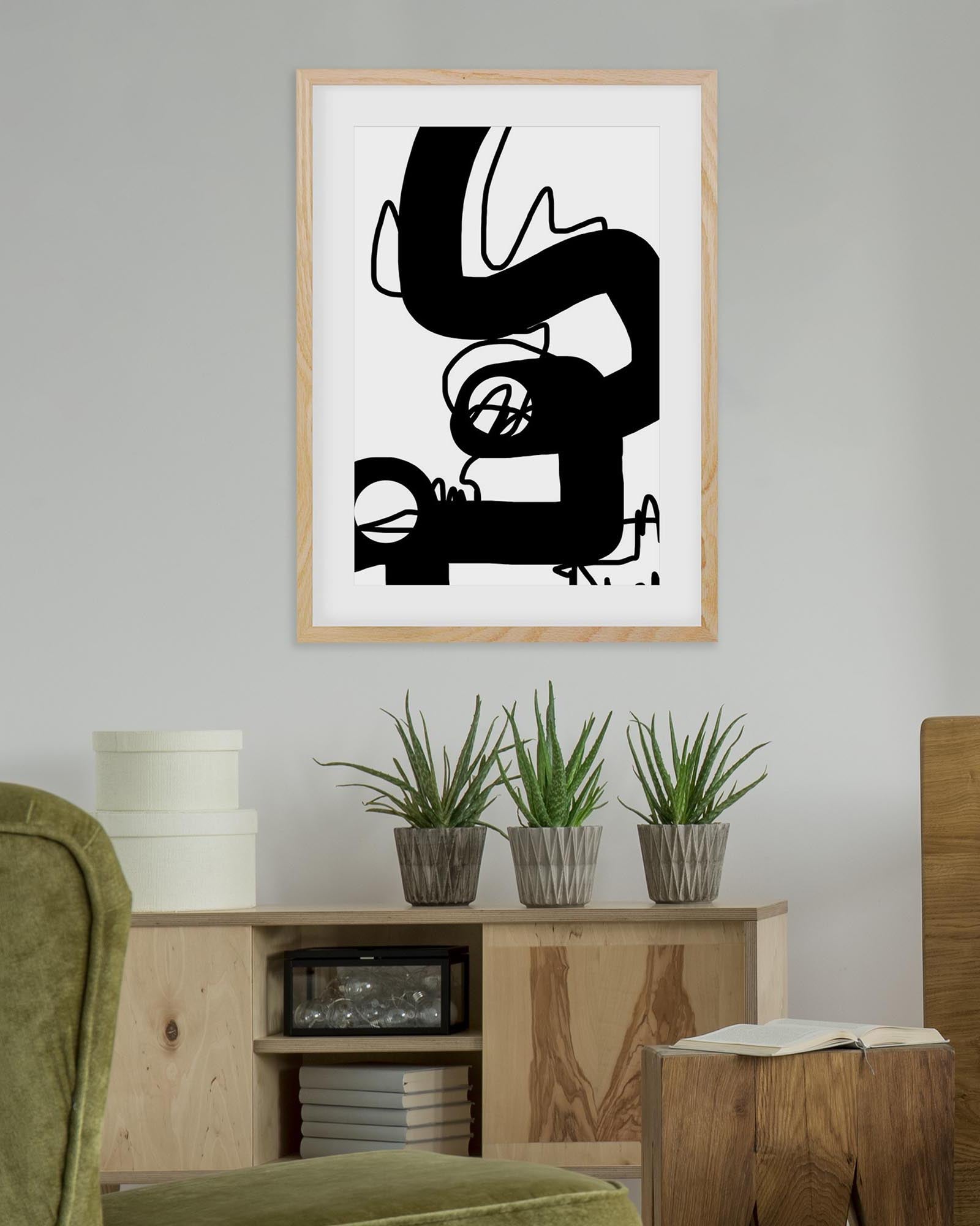 Abstract black and white artwork in a wooden frame on a wall above a wooden cabinet with plants.