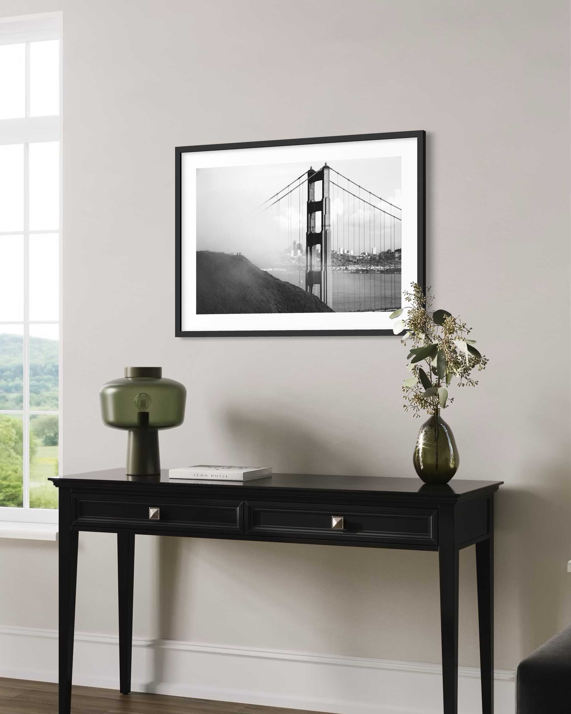 Framed black and white photograph of a suspension bridge on a wall above a black console table with decorative items.