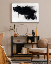 A framed canvas with an abstract black and white design hung on a wall above a console table in a living room setting.
