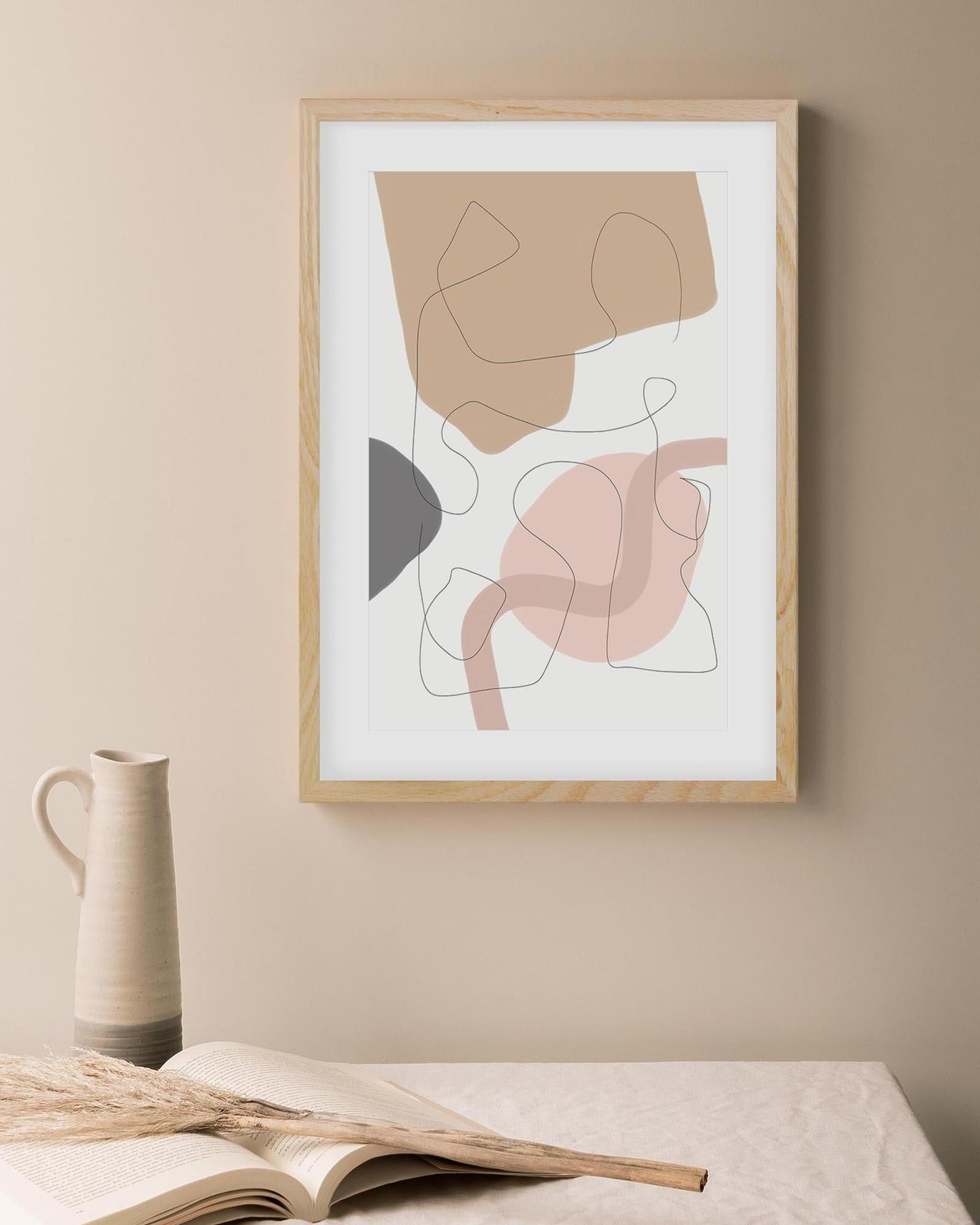 Abstract art print in a wooden frame on a beige wall with a vase and book below.