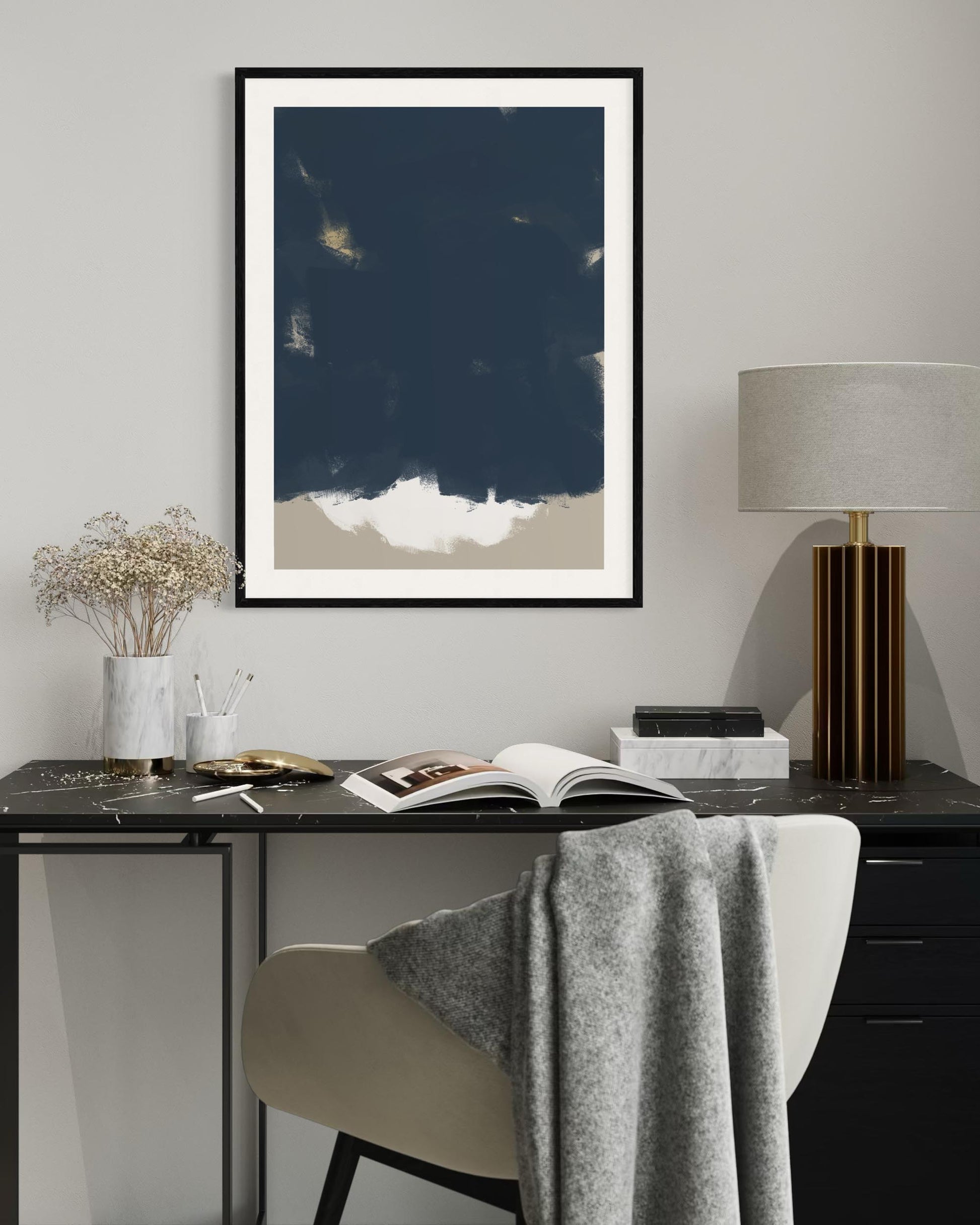 Framed abstract art piece on a wall above a desk with a chair and lamp.